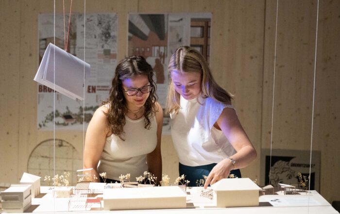 Zwei Frauen stehen an einem Tisch und begutachten Architekturmodelle und Miniaturbauten. Sie wirken konzentriert und kooperativ, während hinter ihnen an der Wand Poster und Entwürfe hängen. at Hochschule Coburg