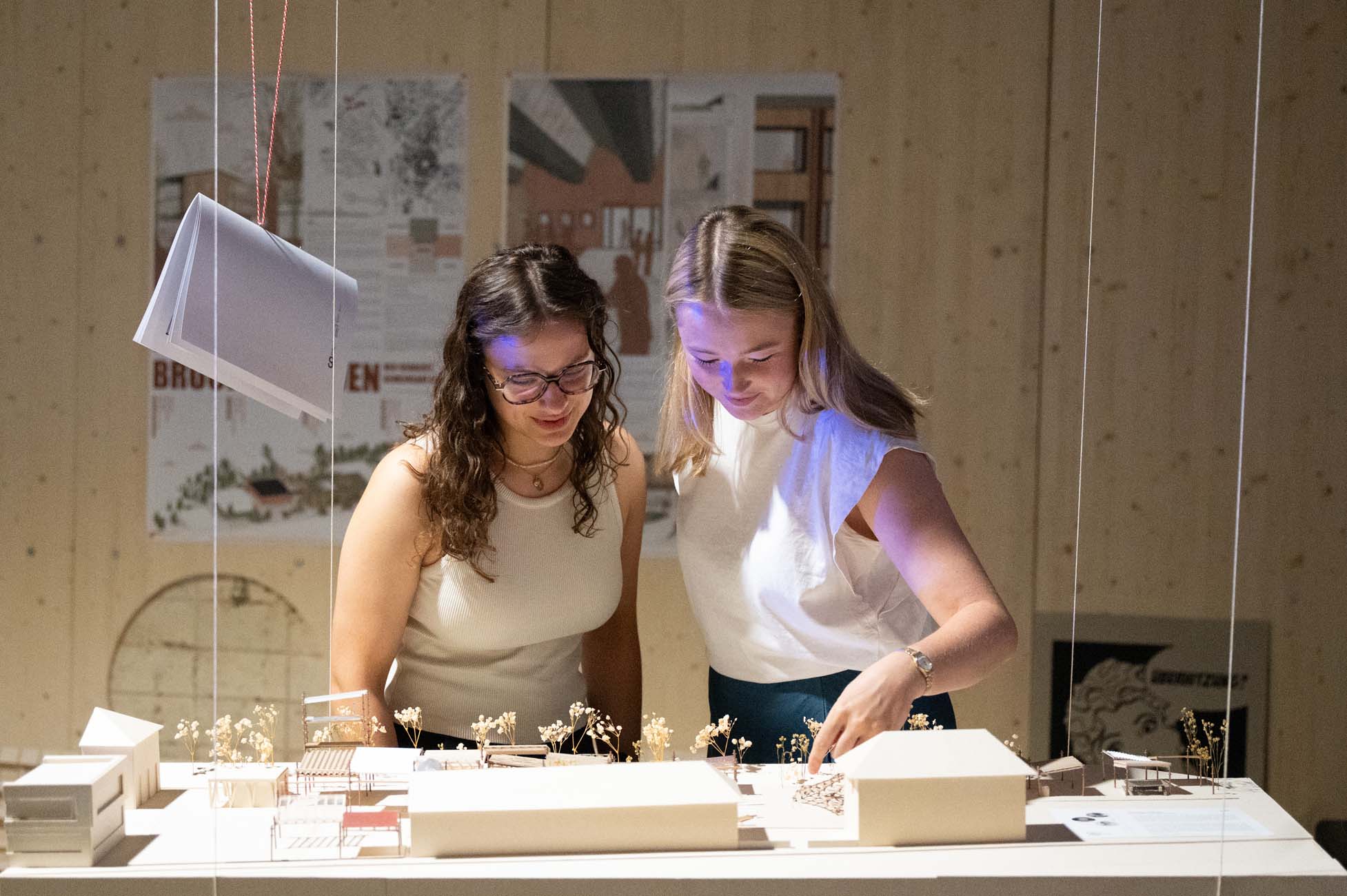 Zwei Frauen stehen an einem Tisch und begutachten Architekturmodelle und Miniaturbauten. Sie wirken konzentriert und kooperativ, während hinter ihnen an der Wand Poster und Entwürfe hängen. at Hochschule Coburg
