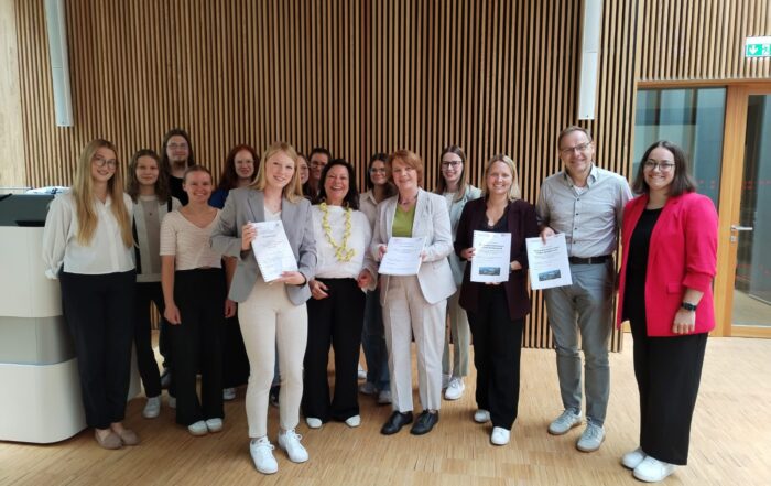 Eine Gruppe von Menschen, meist Frauen, steht in einem Innenraum, hält Urkunden in der Hand und lächelt in die Kamera. Die helle, moderne Umgebung mit Holzwänden unterstreicht ihre Verbindung zum Gesundheitstourismus und zu gesundheitstouristischen Angeboten im Fichtelgebirge. at Hochschule Coburg