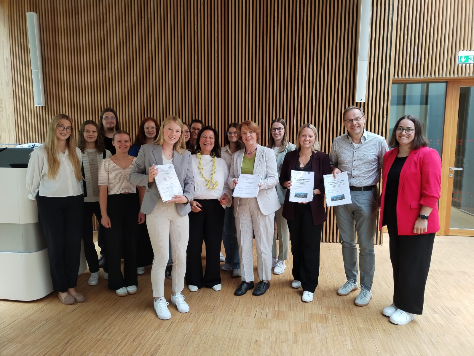 Eine Gruppe von Menschen, meist Frauen, steht in einem Innenraum, hält Urkunden in der Hand und lächelt in die Kamera. Die helle, moderne Umgebung mit Holzwänden unterstreicht ihre Verbindung zum Gesundheitstourismus und zu gesundheitstouristischen Angeboten im Fichtelgebirge. at Hochschule Coburg