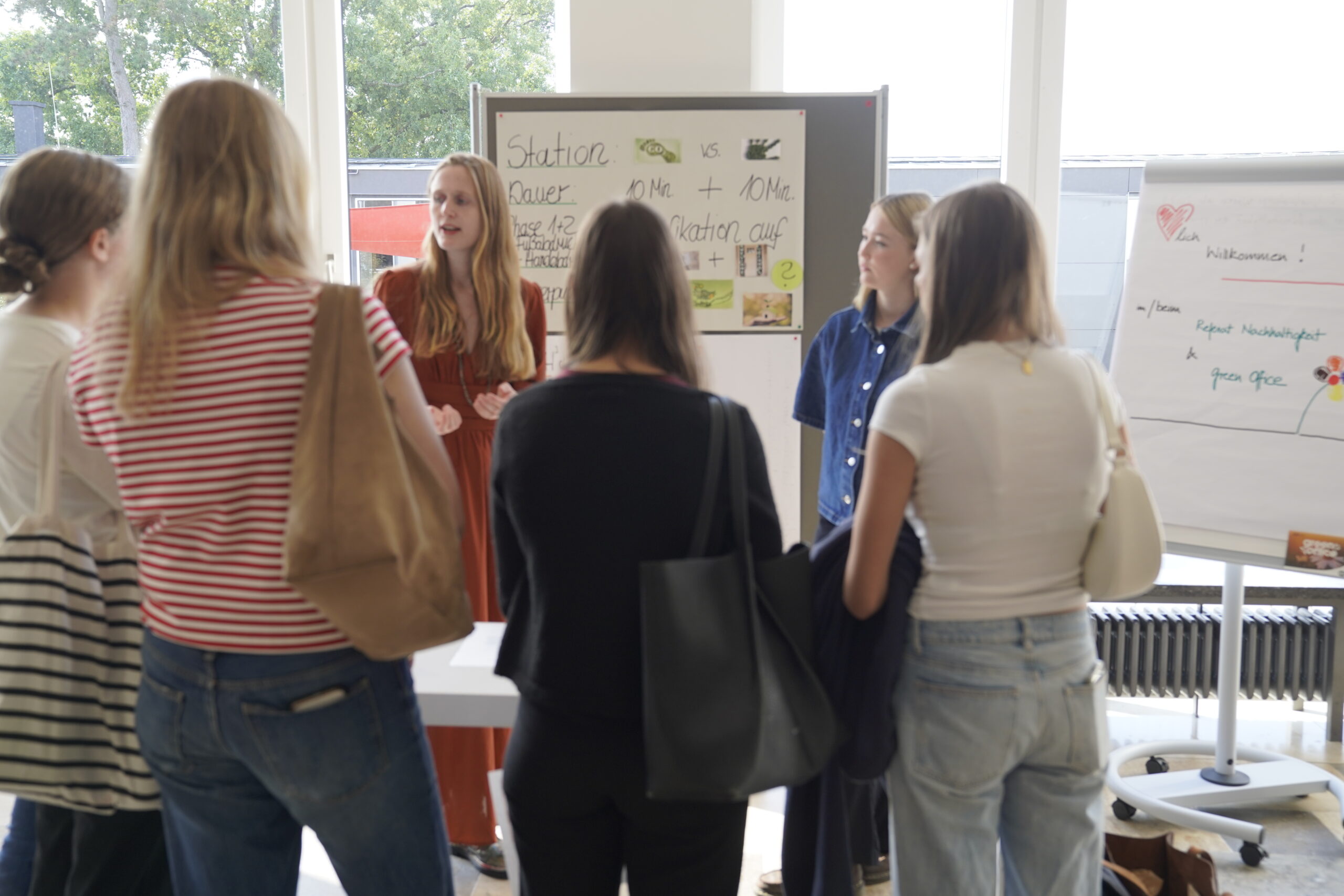 Eine Gruppe von Jugendlichen steht in einem Halbkreis und hört einer Frau zu, die vor einem Whiteboard mit handschriftlichen Notizen und Diagrammen präsentiert - sie erkunden gemeinsam den modernen Raum der Hochschule Coburg. at Hochschule Coburg