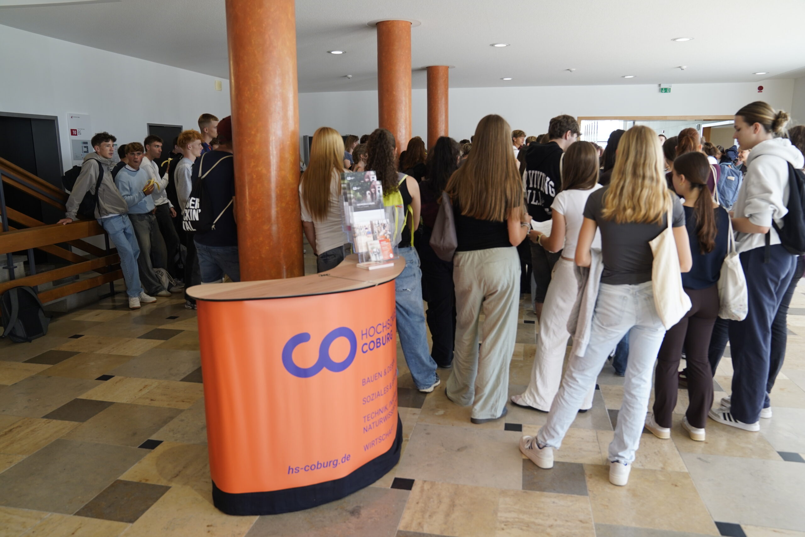 Eine Gruppe von Jugendlichen erkunden die helle Lobby nahe dem orangefarbenen Infoschalter der Hochschule Coburg. Einige stehen in der Schlange, andere unterhalten sich in kleinen Gruppen bei den Fliesen neben der Treppe. at Hochschule Coburg