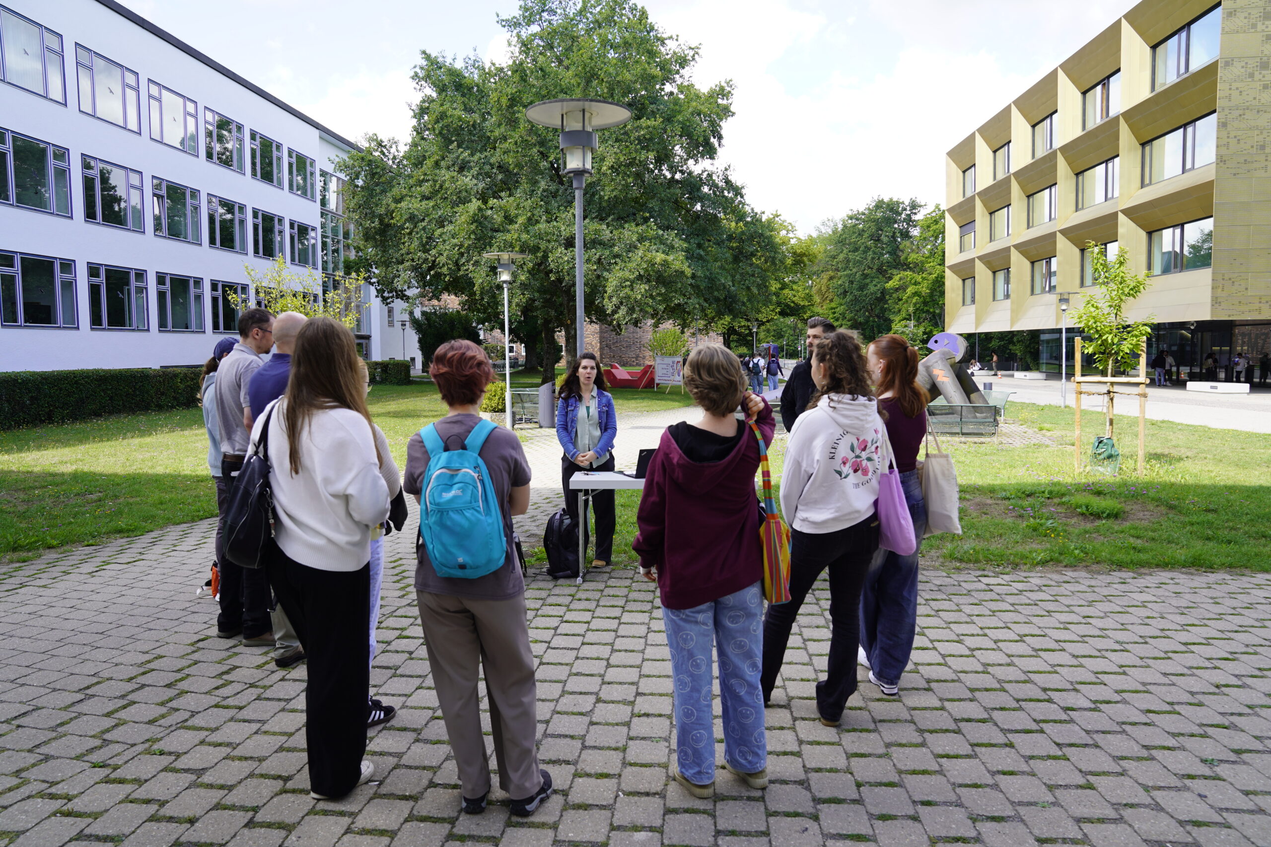 Eine Gruppe Jugendlicher steht im Halbkreis um einen kleinen Tisch im Freien auf einem gepflasterten Gehweg, hinter dem zwei Personen Platz nehmen. Im Hintergrund sind moderne Gebäude und grüne Bäume der Hochschule Coburg zu sehen, während sie die Umgebung erkunden. at Hochschule Coburg