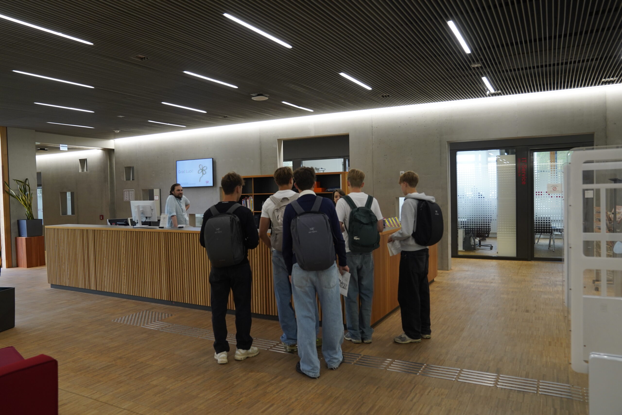 Fünf Jugendliche mit Rucksäcken erkunden das offene, lichtdurchflutete Foyer der Hochschule Coburg und sprechen am modernen Empfangstresen mit einer Mitarbeiterin. at Hochschule Coburg