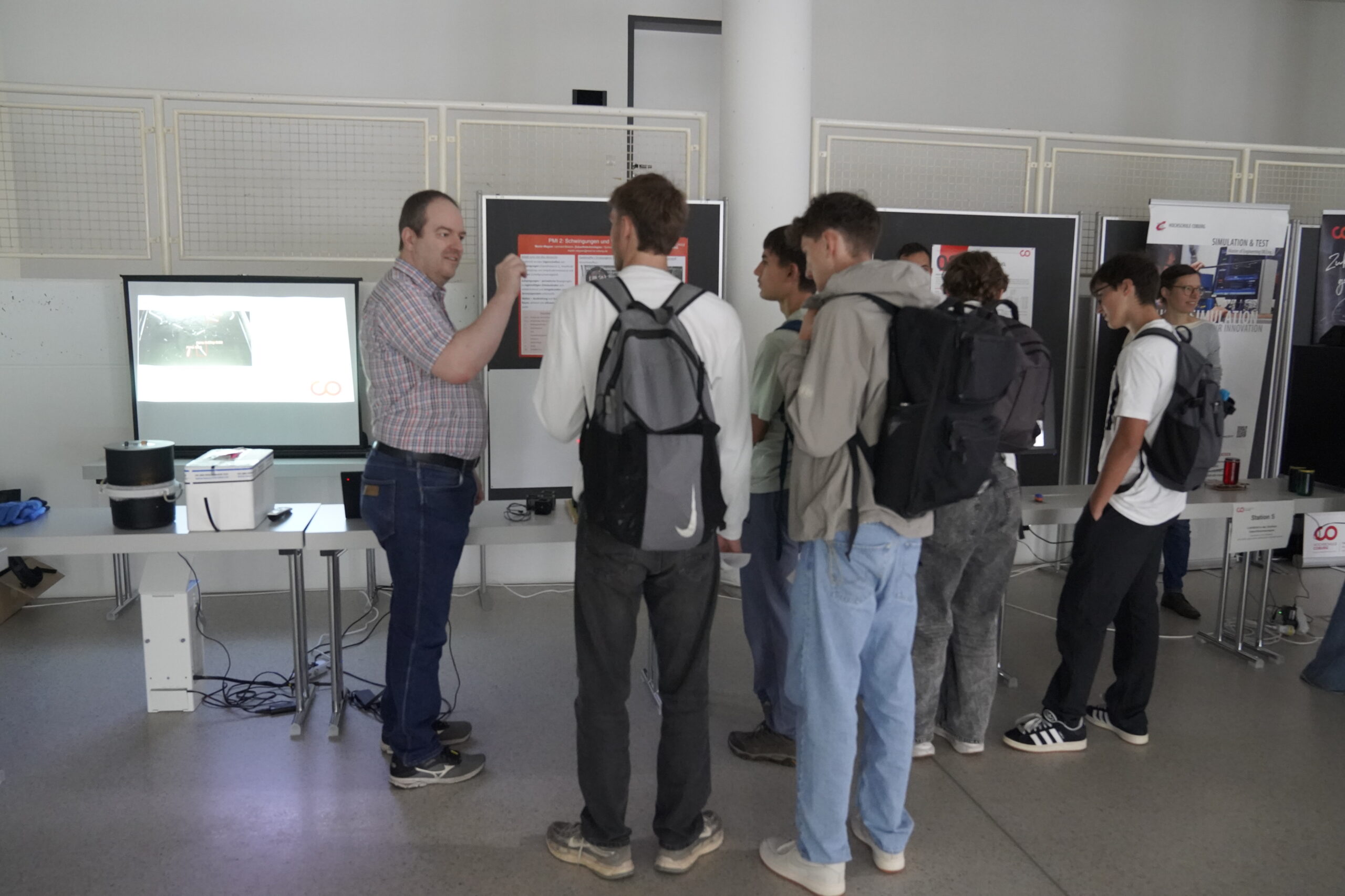 Ein Mann erklärt einer Gruppe von Jugendlichen mit Rucksäcken Informationen auf einem Bildschirm, während sie eine Indoor-Veranstaltung an der Hochschule Coburg erkunden. An der Wand hinter ihnen sind mehrere Bildschirme und Plakate aufgestellt. at Hochschule Coburg