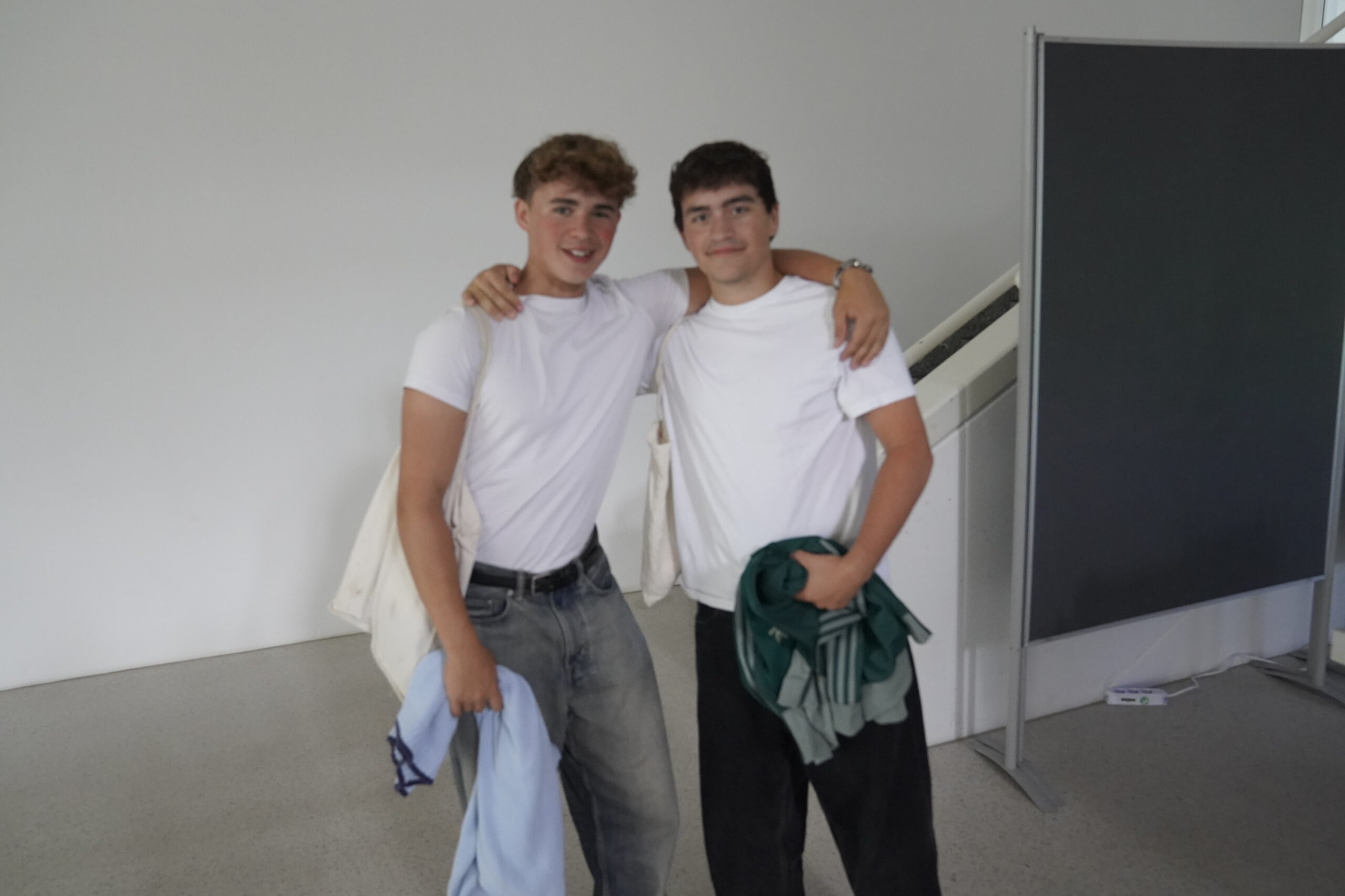 Zwei Jugendliche stehen in einem Haus und halten sich in den Armen, beide tragen weiße T-Shirts. Einer hält eine blaue Jacke, der andere grüne Kleidung. Sie lächeln, und im Hintergrund ist eine Treppe zu sehen, möglicherweise in der Hochschule Coburg. at Hochschule Coburg