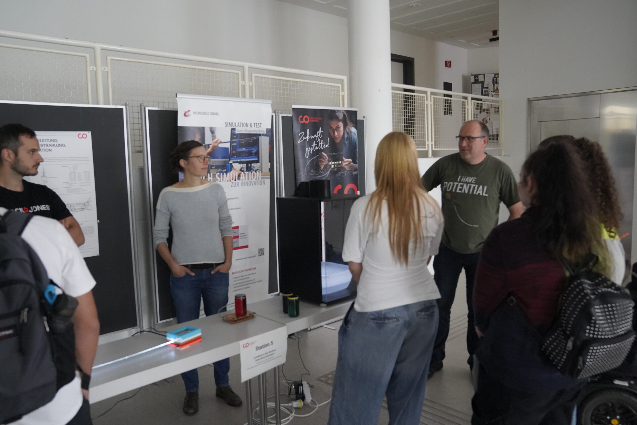 Eine Gruppe Jugendlicher steht um einen Tisch mit Schautafeln und Informationsplakaten herum und unterhält sich, während sie innovative Projekte bei einer Veranstaltung der Hochschule Coburg im Innenbereich erkunden. at Hochschule Coburg