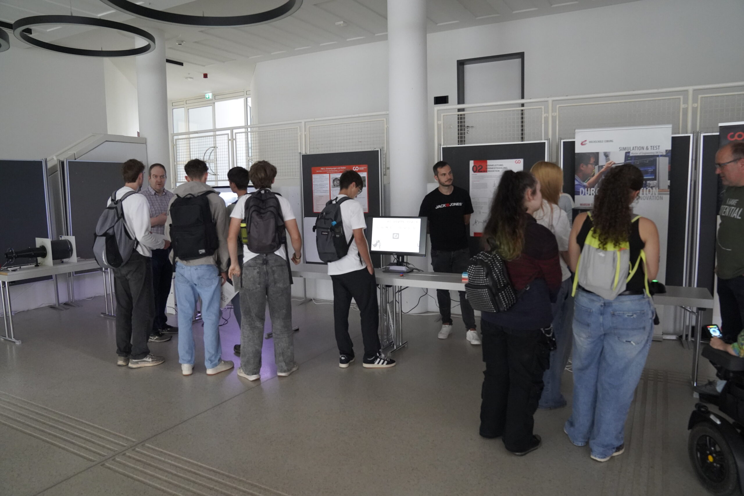 Eine Gruppe von Jugendlichen mit Rucksäcken erkunden Ausstellungstische mit Monitoren und Postern in einem modernen Innenraum der Hochschule Coburg mit grauen Böden und weißen Wänden. at Hochschule Coburg