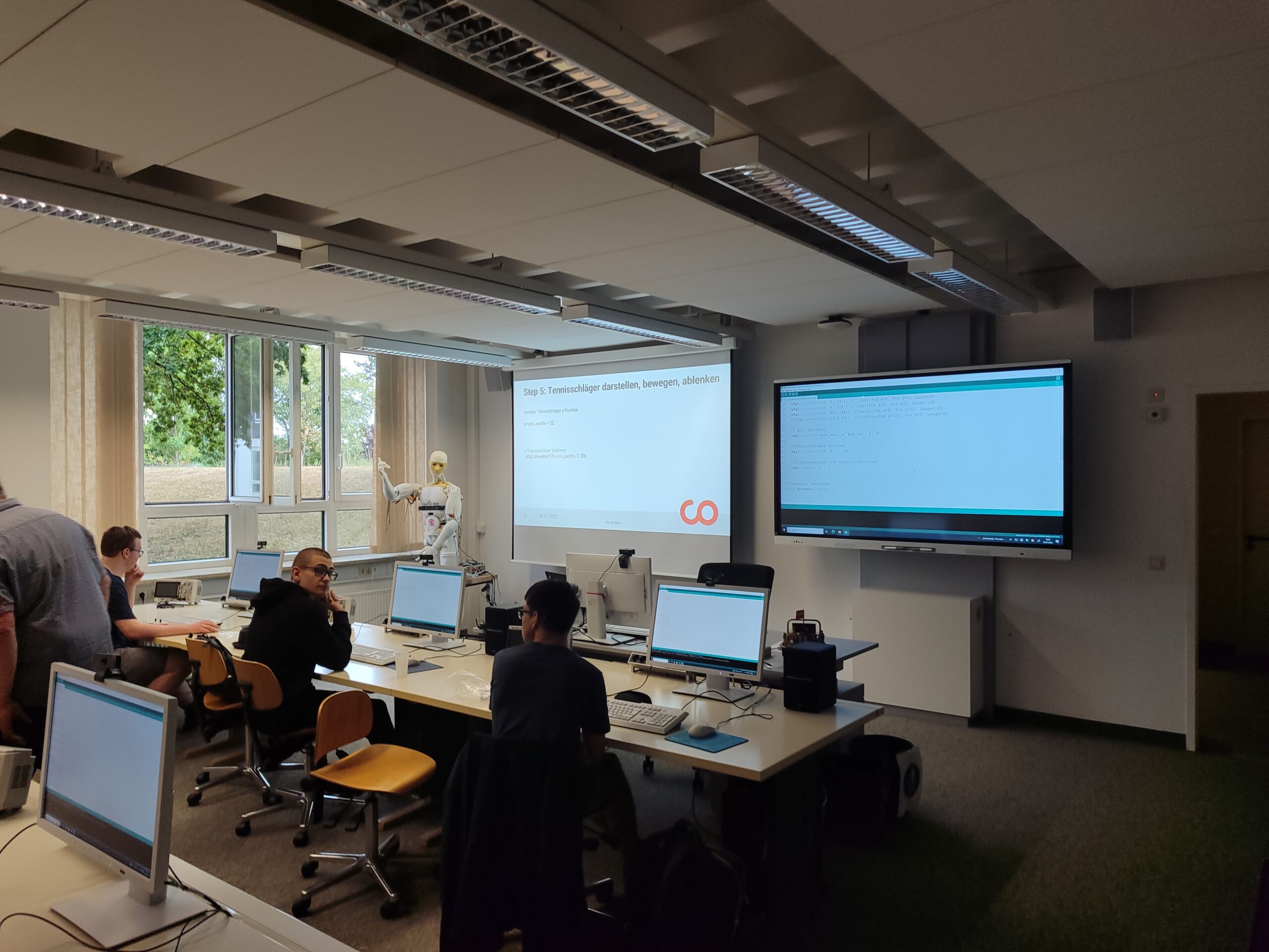 Ein modernes Klassenzimmer mit Schülern an Schreibtischen, die mit Computern arbeiten, einem Lehrer, der an einer Projektionsfläche eine Präsentation zeigt, und einem großen Monitor, der einen Code anzeigt. Natürliches Licht strömt herein, und auf dem Bildschirm erscheint ein automatisch gespeicherter Entwurf. at Hochschule Coburg