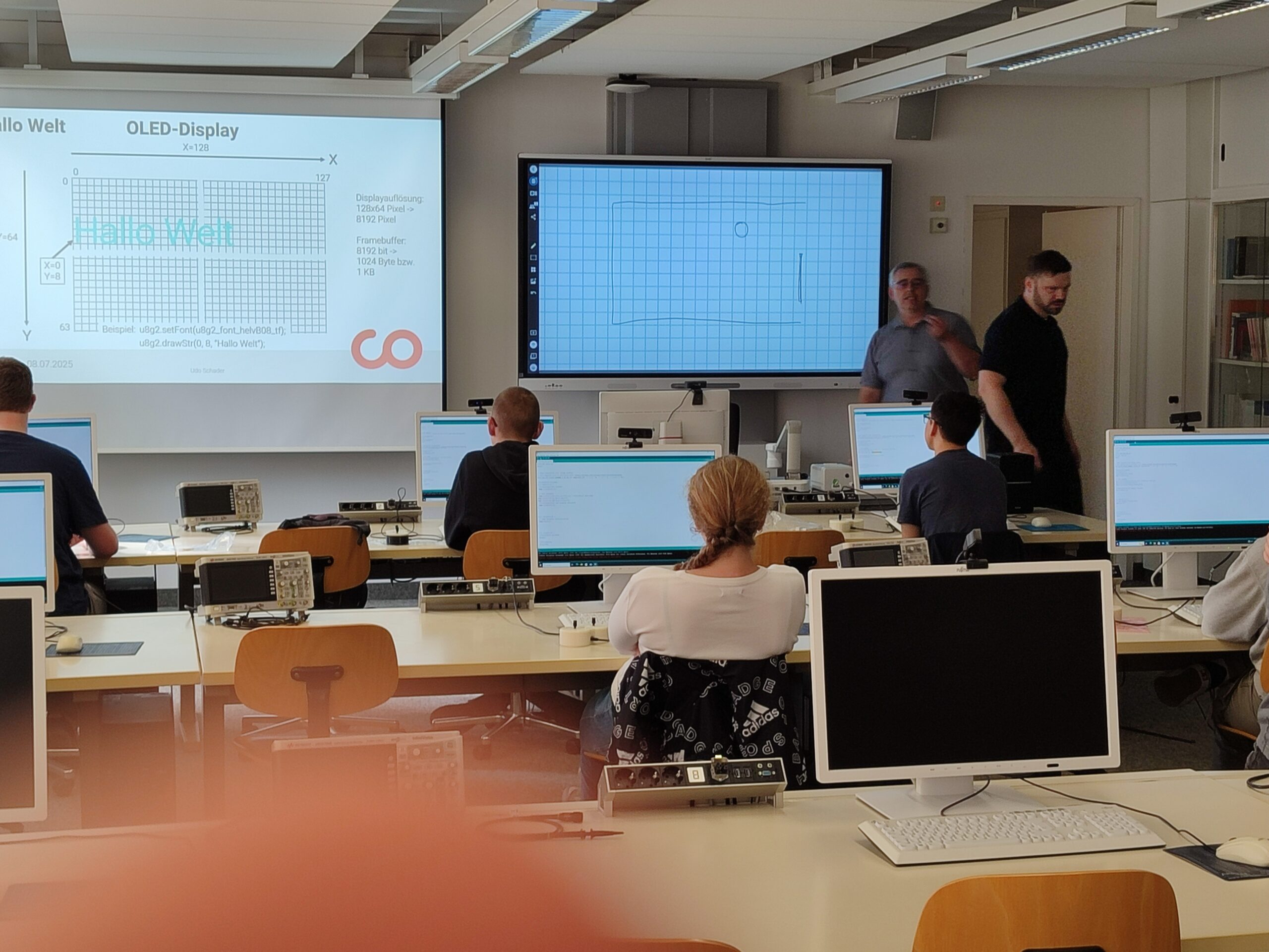 Schüler sitzen in einem Klassenzimmer an Tischen mit Computern und blicken auf einen großen Bildschirm, auf dem ein OLED-Displaydiagramm und "Hello Welt" zu sehen ist. Zwei Lehrkräfte stehen am Whiteboard und diskutieren einen automatisch gespeicherten Entwurf. at Hochschule Coburg