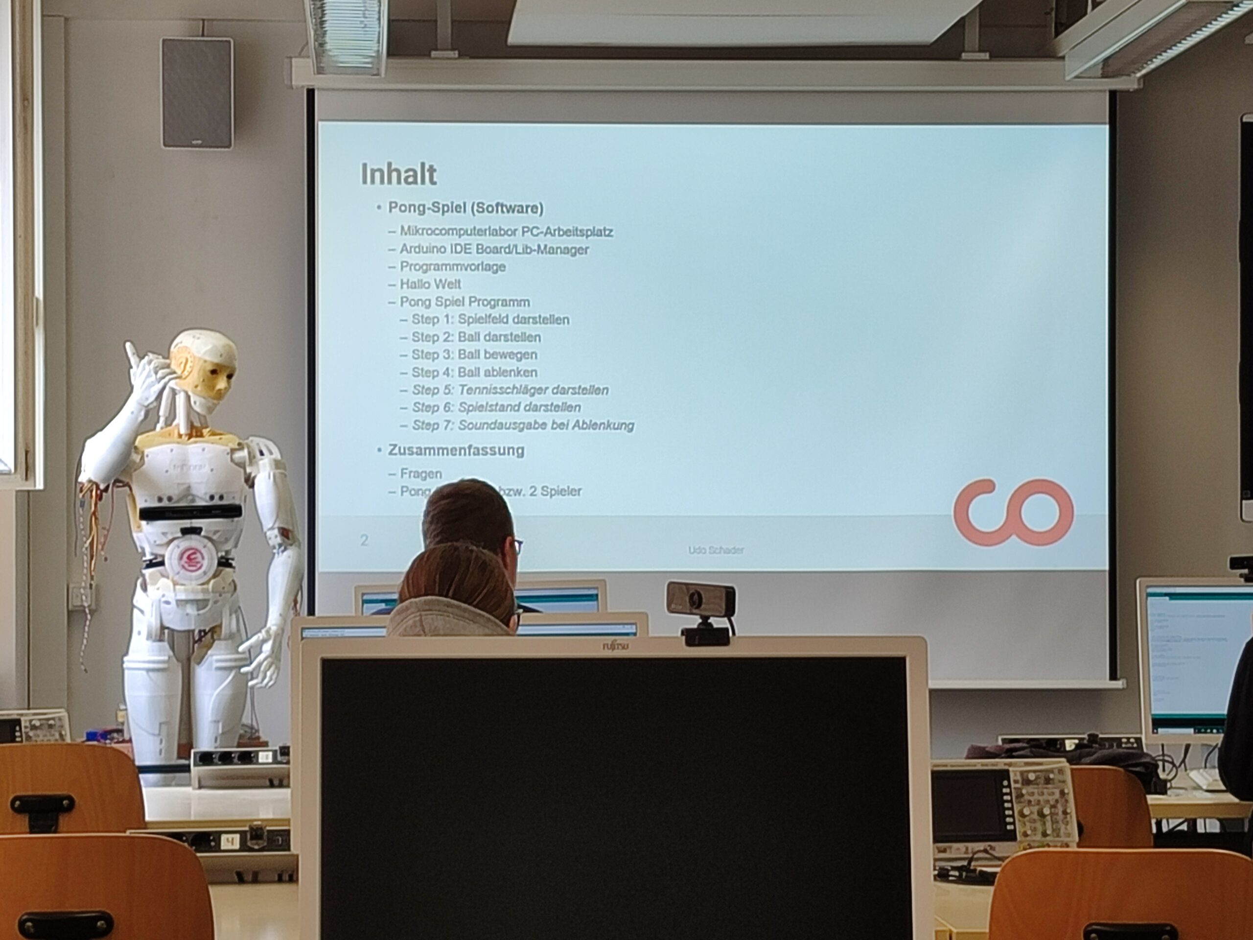 Ein Klassenzimmer mit Schülern, die vor einem Bildschirm sitzen, auf dem eine Präsentation über die Programmierung eines Pong-Spiels gezeigt wird. Auf der linken Seite steht ein humanoider Roboter, und im Vordergrund sitzt eine Person an einem Computer und arbeitet an einem automatisch gespeicherten Entwurf. at Hochschule Coburg