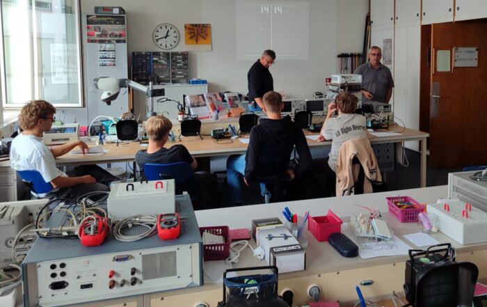 Ein Klassenzimmer mit Schülern, die an Tischen sitzen und mit elektronischen Geräten arbeiten, während zwei Lehrer in der Nähe einer Tafel im vorderen Teil des Raums stehen. Auf den Tischen und in den Regalen sind verschiedene Werkzeuge und Geräte zu sehen, darunter ein automatisch gespeicherter Entwurf. at Hochschule Coburg