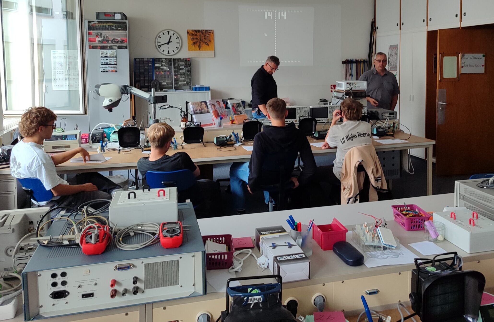 Ein Klassenzimmer mit Schülern, die an Tischen sitzen und mit elektronischen Geräten arbeiten, während zwei Lehrer in der Nähe einer Tafel im vorderen Teil des Raums stehen. Auf den Tischen und in den Regalen sind verschiedene Werkzeuge und Geräte zu sehen, darunter ein automatisch gespeicherter Entwurf. at Hochschule Coburg