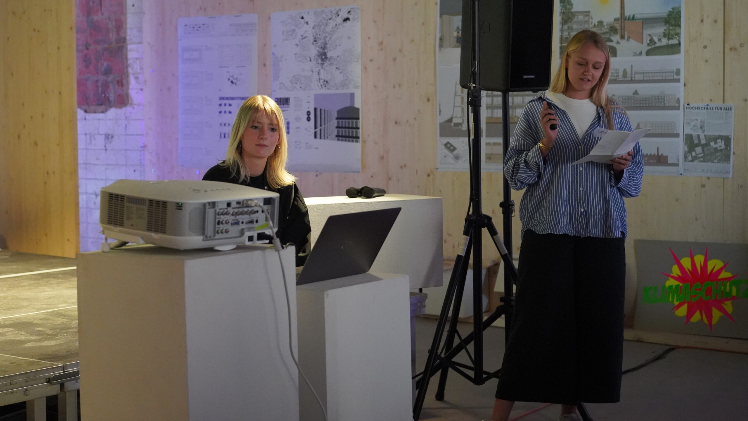 Zwei Frauen halten in einem Innenraum eine Präsentation; die eine sitzt an einem Laptop neben einem Projektor, die andere steht mit einem Mikrofon in der Hand und liest von einem Blatt Papier ab. An der Wand hängen Architekturplakate, die auf nachhaltige Stadtentwicklung hinweisen. at Hochschule Coburg