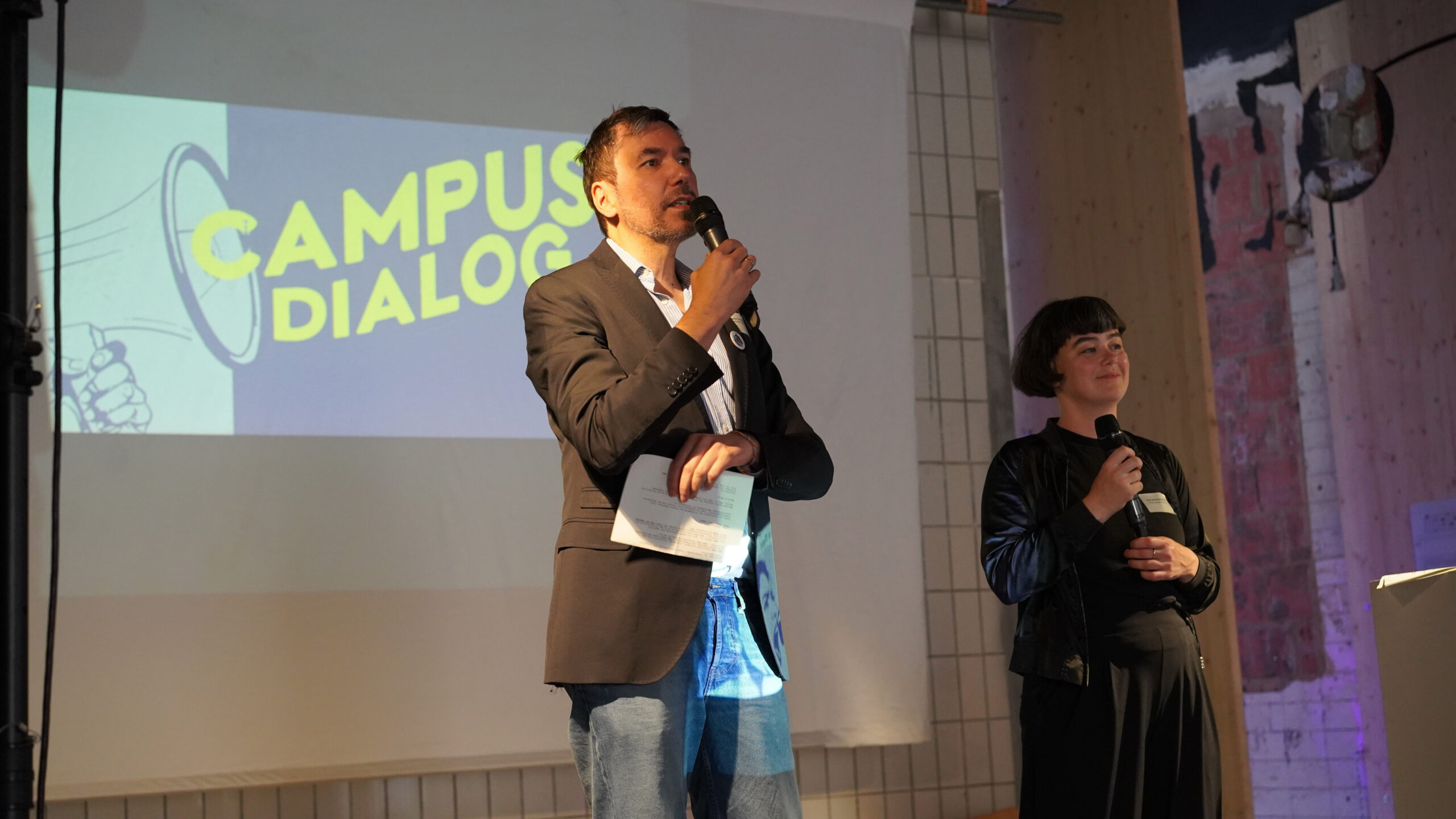 Ein Mann und eine Frau stehen auf der Bühne und halten Mikrofone. Hinter ihnen zeigt ein Bildschirm eine Megaphon-Grafik und die Worte "CAMPUS DIALOG", die auf Diskussionen über transformative Stadtentwicklung in einem Veranstaltungsraum hinweisen. at Hochschule Coburg