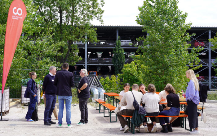 Eine Gruppe von Menschen hat sich im Freien an orangefarbenen Bänken und Tischen versammelt und führt eine lebhafte Diskussion über transformative Stadtentwicklung. Grüne Bäume und ein mehrstöckiges Parkhaus dienen als Kulisse für ihre Gespräche. at Hochschule Coburg