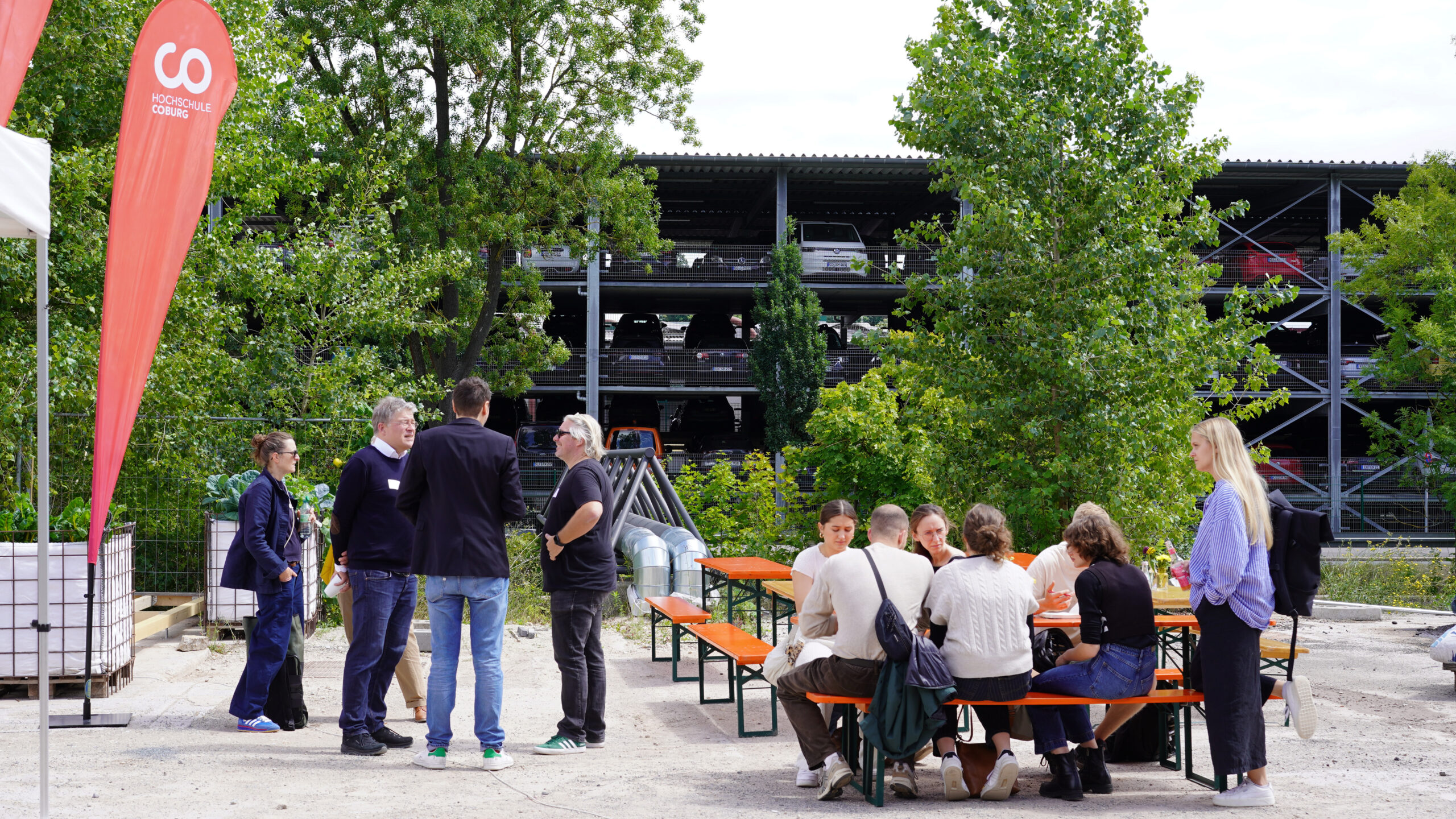 Eine Gruppe von Menschen hat sich im Freien an orangefarbenen Bänken und Tischen versammelt und führt eine lebhafte Diskussion über transformative Stadtentwicklung. Grüne Bäume und ein mehrstöckiges Parkhaus dienen als Kulisse für ihre Gespräche. at Hochschule Coburg