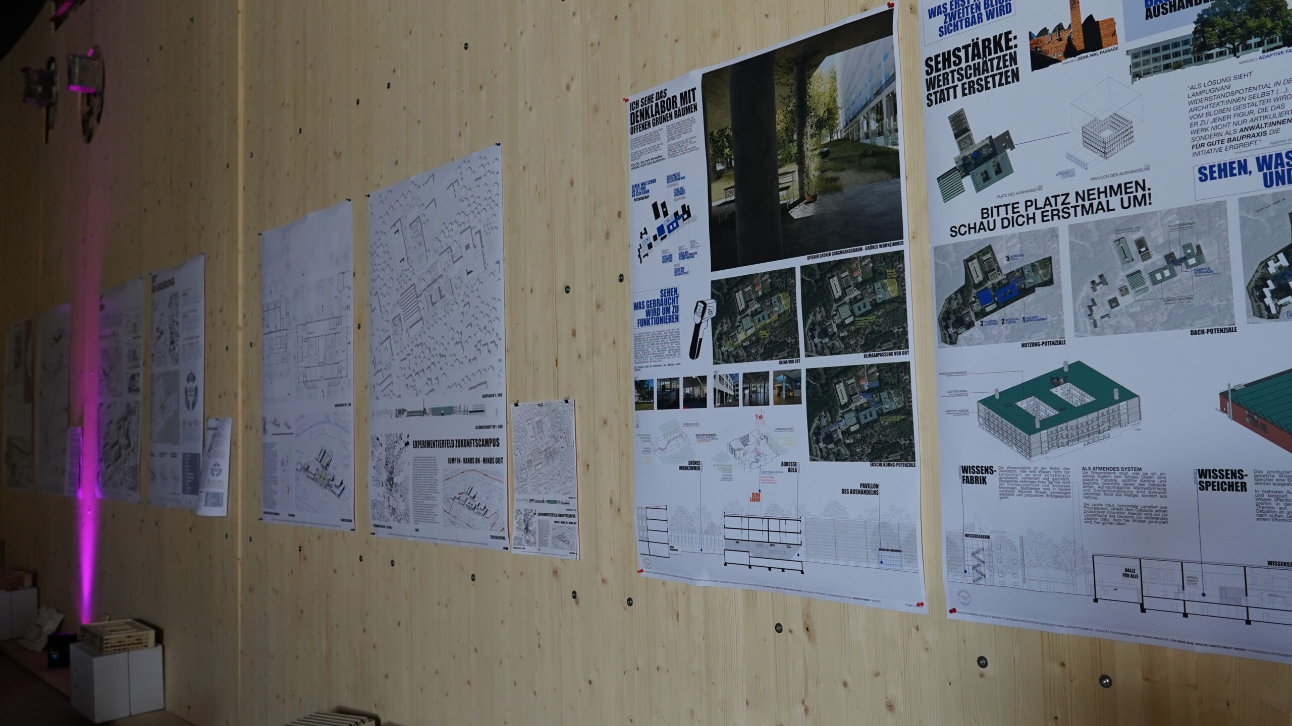 An einer hellen Holzwand sind mehrere Architekturplakate und technische Zeichnungen angebracht, die sich mit der transformativen Stadtentwicklung befassen. Auf den Postern sind Diagramme, Karten, Baupläne und Texte abgebildet, wobei die Abschnitte in Blau und Grün hervorgehoben sind. at Hochschule Coburg
