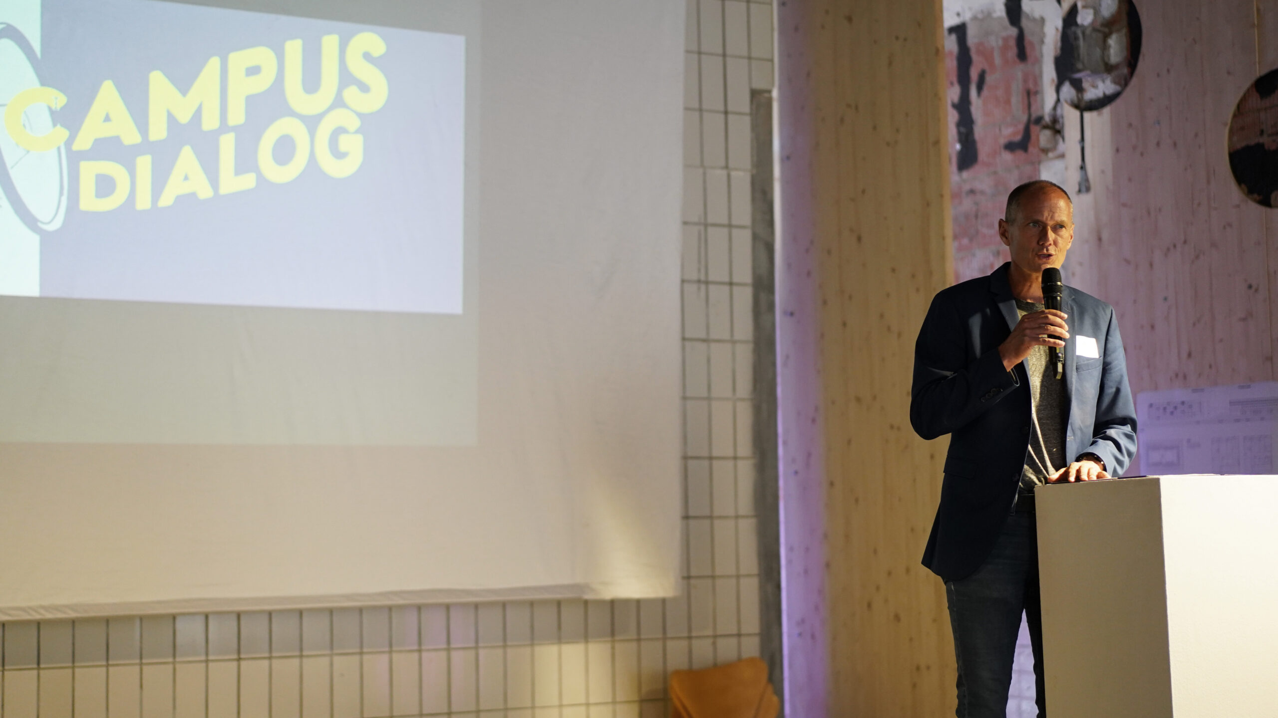 Ein Mann in einem Blazer spricht von einem Podium aus in ein Mikrofon. Hinter ihm wird eine Folie mit der Aufschrift "CAMPUS DIALOG" projiziert, auf der Themen wie transformative Stadtentwicklung und Ideen für eine nachhaltige urbane Zukunft dargestellt werden. at Hochschule Coburg