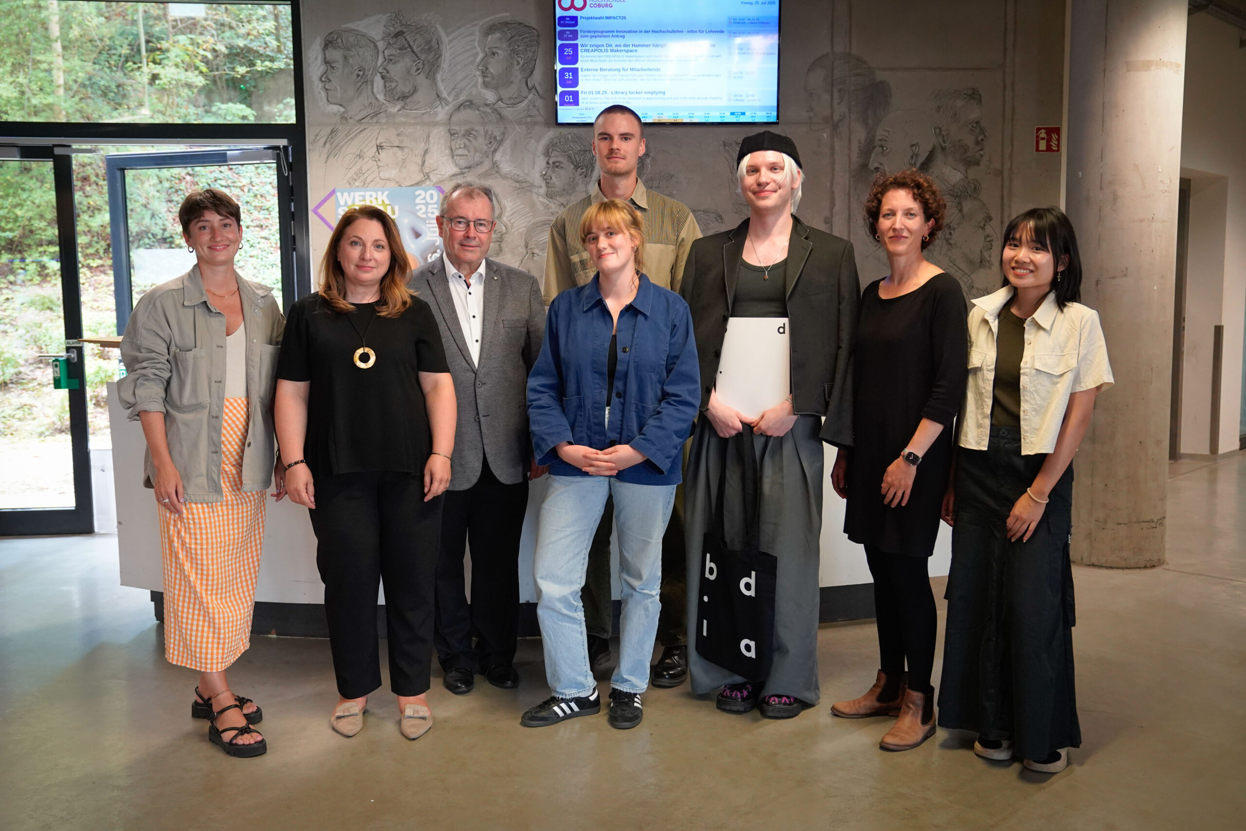 Acht Menschen stehen gemeinsam in einem Innenraum vor einer Wand mit künstlerischen Gravuren und einem digitalen Bildschirm und posieren für ein Gruppenfoto. Als Teil der bdia-Veranstaltung feiern sie Abschlussarbeiten, die Tabus brechen im Design. at Hochschule Coburg