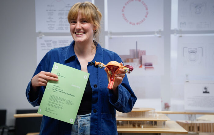 Eine lächelnde Person in einer blauen Jacke hält ein grünes Blatt Papier mit der Aufschrift "Abschlussarbeiten" in der einen und ein Modell des weiblichen Fortpflanzungssystems in der anderen Hand und steht vor Schautafeln, die Tabus brechen mit informativen Diagrammen und Skizzen helfen. at Hochschule Coburg