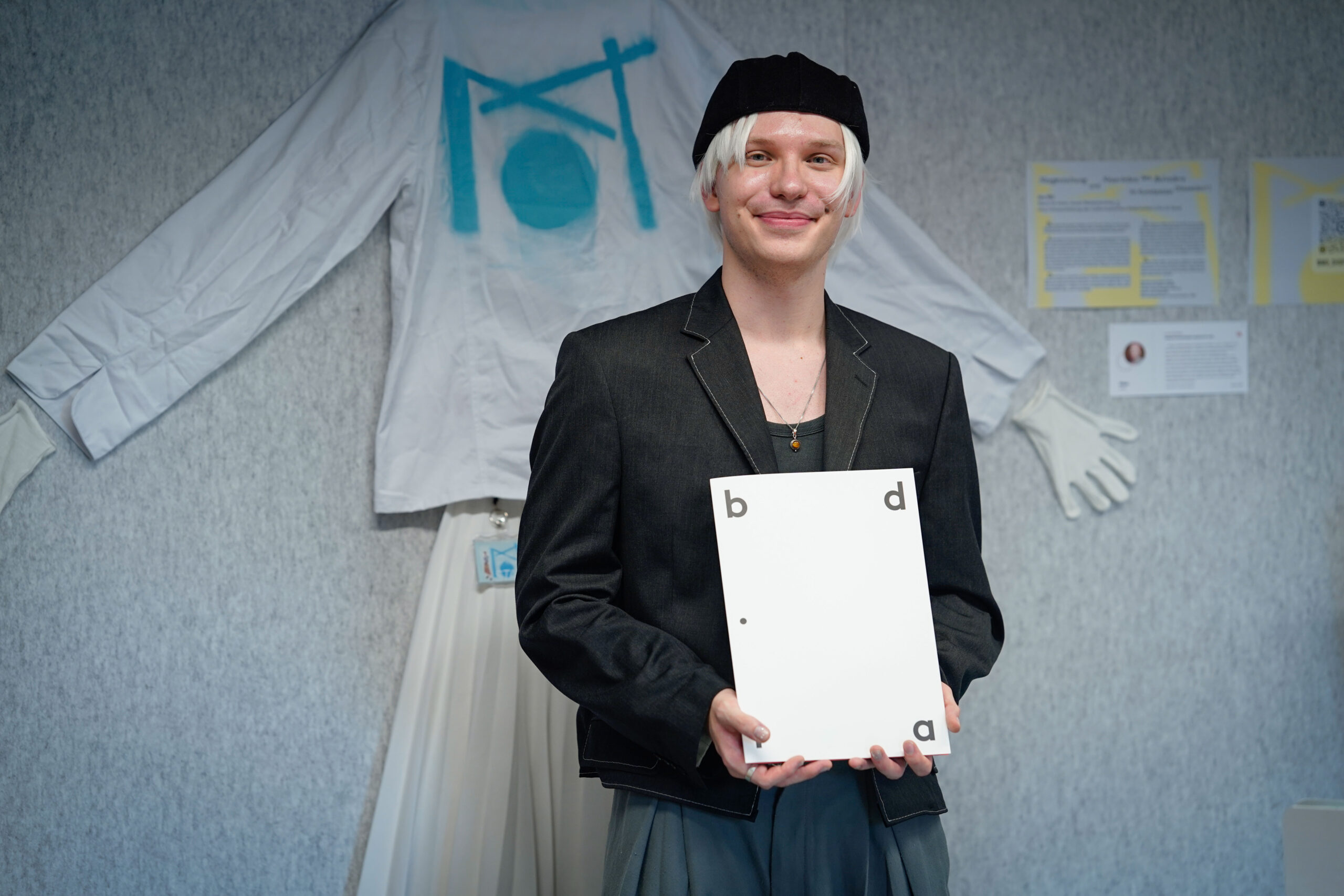 Eine Person, die einen schwarzen Blazer, eine graue Baskenmütze und eine weiße Perücke trägt, lächelt und hält eine weiße Tafel mit den Kleinbuchstaben "b", "d" und "a", die auf bdiα verweisen. Dahinter ist ein großes weißes Hemd mit blauen Motiven zu sehen, die Tabus brechen in Abschlussarbeiten widerspiegeln. at Hochschule Coburg