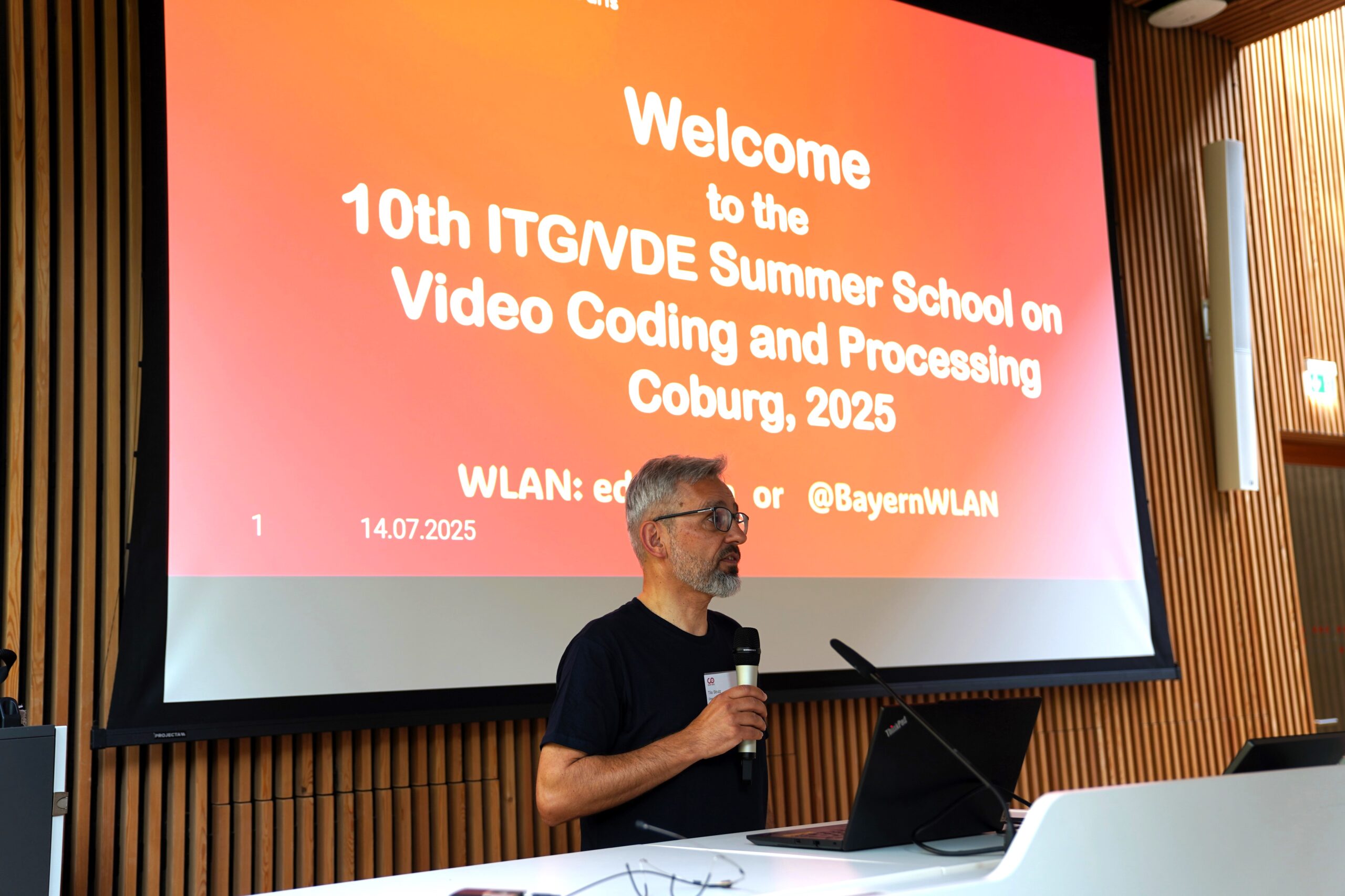 Ein Mann mit grauen Haaren und Brille spricht in ein Mikrofon auf der Bühne vor einem großen Bildschirm mit der Aufschrift "Welcome to the 10th ITG/VDE Summer School on Video Coding and Processing Coburg, 2025" und hebt die innovative Forschung hervor. at Hochschule Coburg