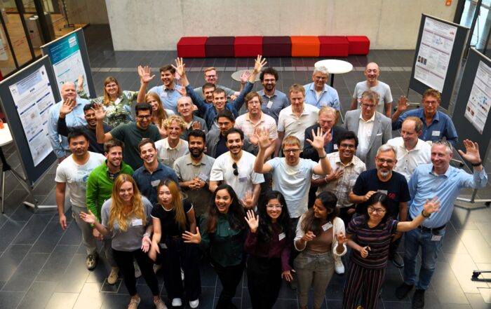 Eine bunt gemischte Gruppe von Menschen steht in einem Innenraum, lächelt und winkt in die Kamera vor wissenschaftlichen Postern während einer Summer School-Veranstaltung, mit roten und orangefarbenen Sitzgelegenheiten im Hintergrund. Die Atmosphäre ist lebhaft und freundlich. at Hochschule Coburg