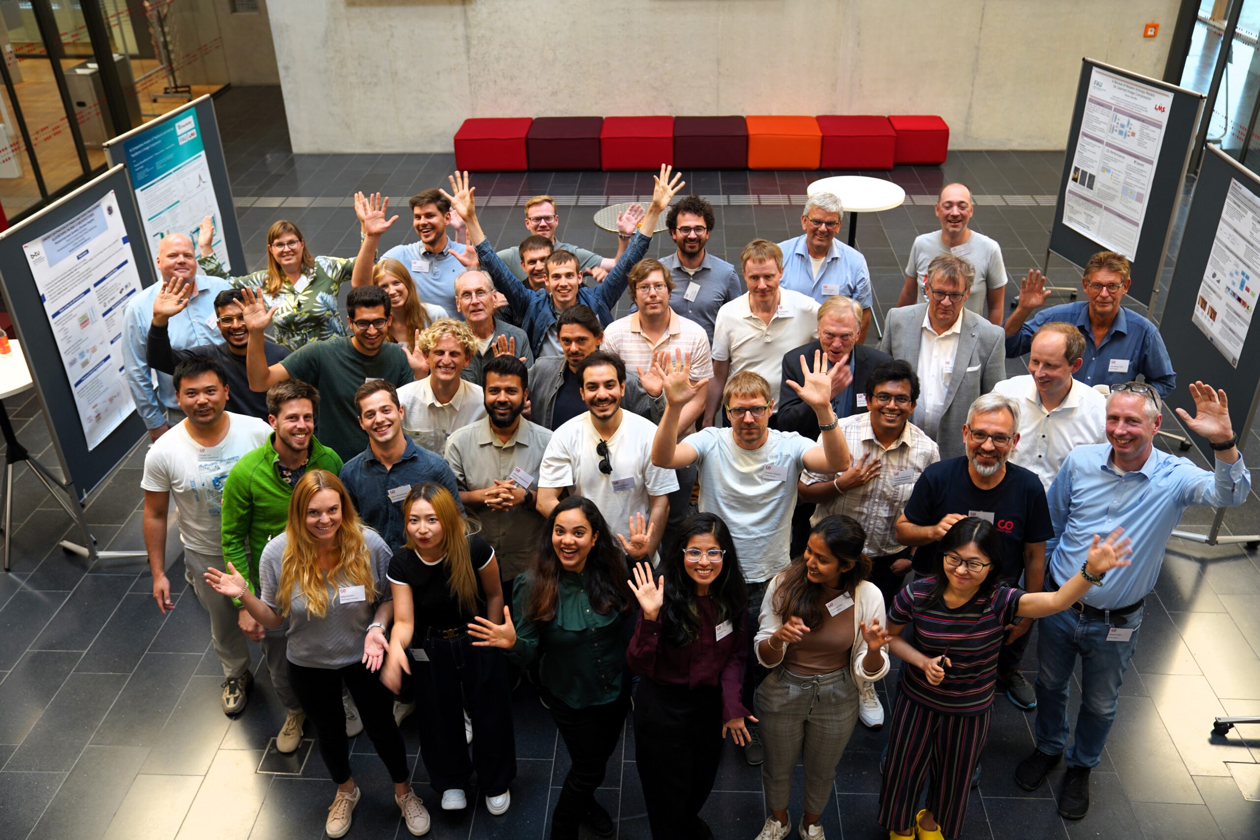 Eine bunt gemischte Gruppe von Menschen steht in einem Innenraum, lächelt und winkt in die Kamera vor wissenschaftlichen Postern während einer Summer School-Veranstaltung, mit roten und orangefarbenen Sitzgelegenheiten im Hintergrund. Die Atmosphäre ist lebhaft und freundlich. at Hochschule Coburg