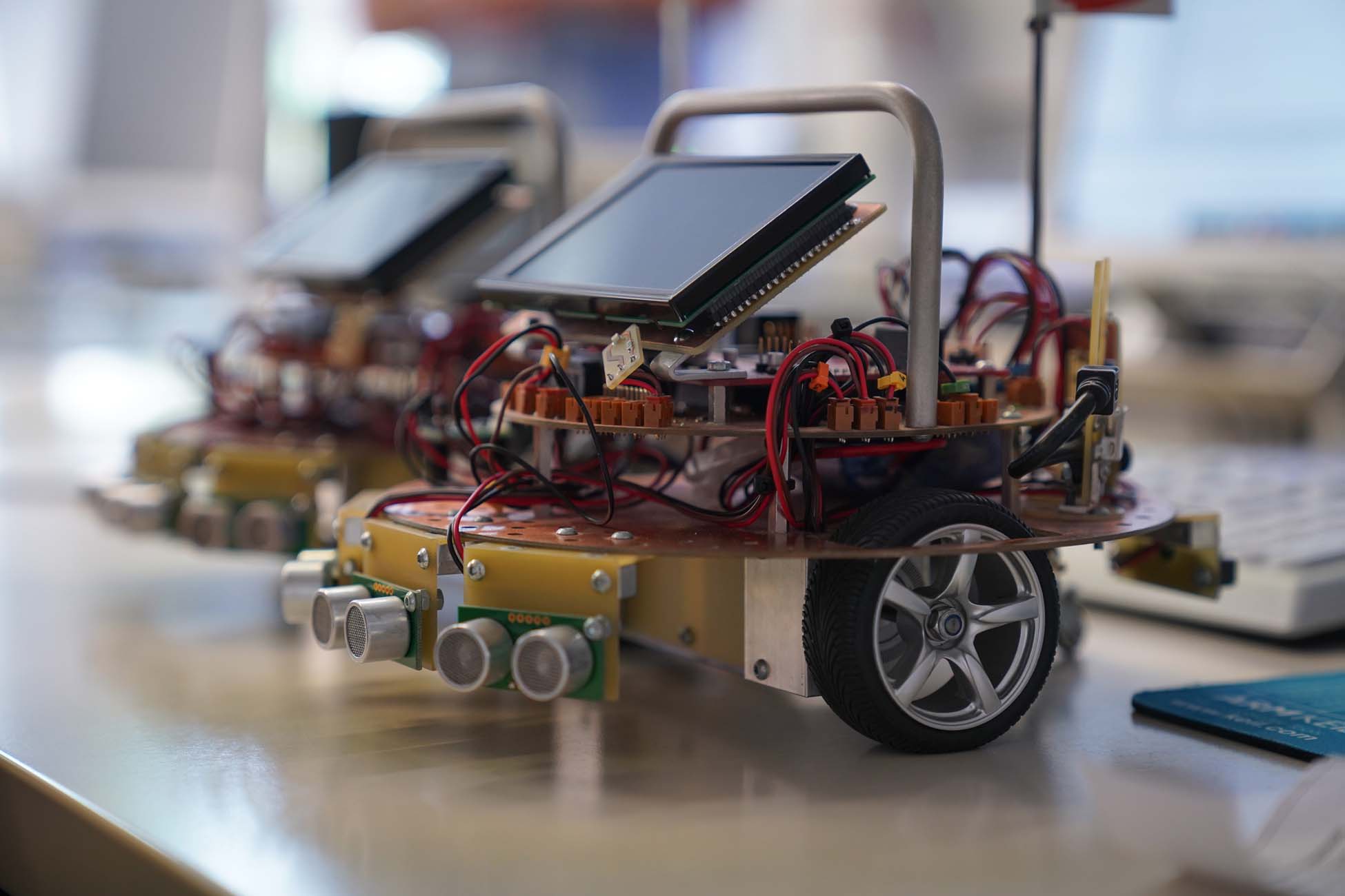 Ein kleiner Radroboter mit freiliegenden Platinen, Drähten, Sensoren und einem Bildschirm steht auf einem Tisch an der Hochschule Coburg und lädt Jugendliche zum Erkunden ein, während ein anderer ähnlicher Roboter unscharf im Hintergrund zu sehen ist. at Hochschule Coburg