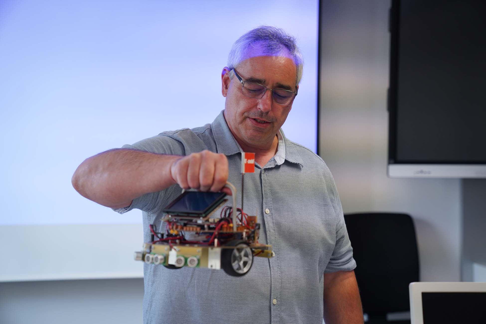 Ein Mann mit grauen Haaren und Brille hält einen kleinen Roboter auf Rädern mit Drähten und elektronischen Bauteilen in einem Klassenzimmer der Hochschule Coburg, wo Jugendliche innovative Technik erkunden. at Hochschule Coburg