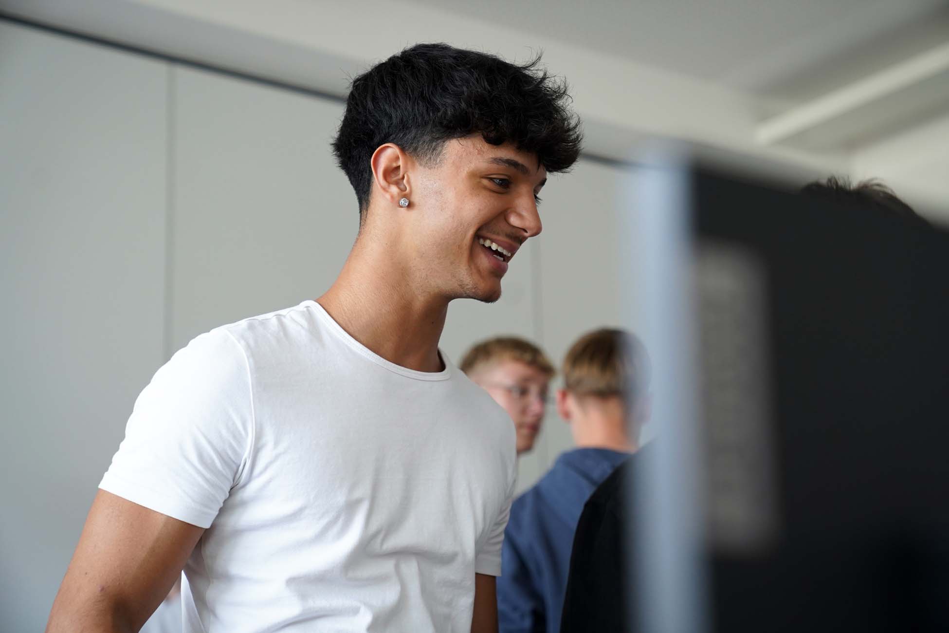Ein junger Mann in einem weißen T-Shirt lächelt beim Blick auf einen Monitor, während drei andere Jugendliche im Hintergrund bei der Hochschule Coburg erkunden. at Hochschule Coburg