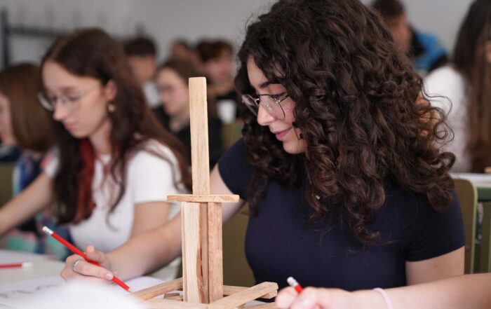 Eine Jugendliche mit lockigem Haar und Brille studiert an ihrem Schreibtisch eine Holzstruktur mit einem roten Bleistift, während sie von anderen Studierenden der Hochschule Coburg im Klassenraum umgeben ist und gemeinsam erkunden lernt. at Hochschule Coburg