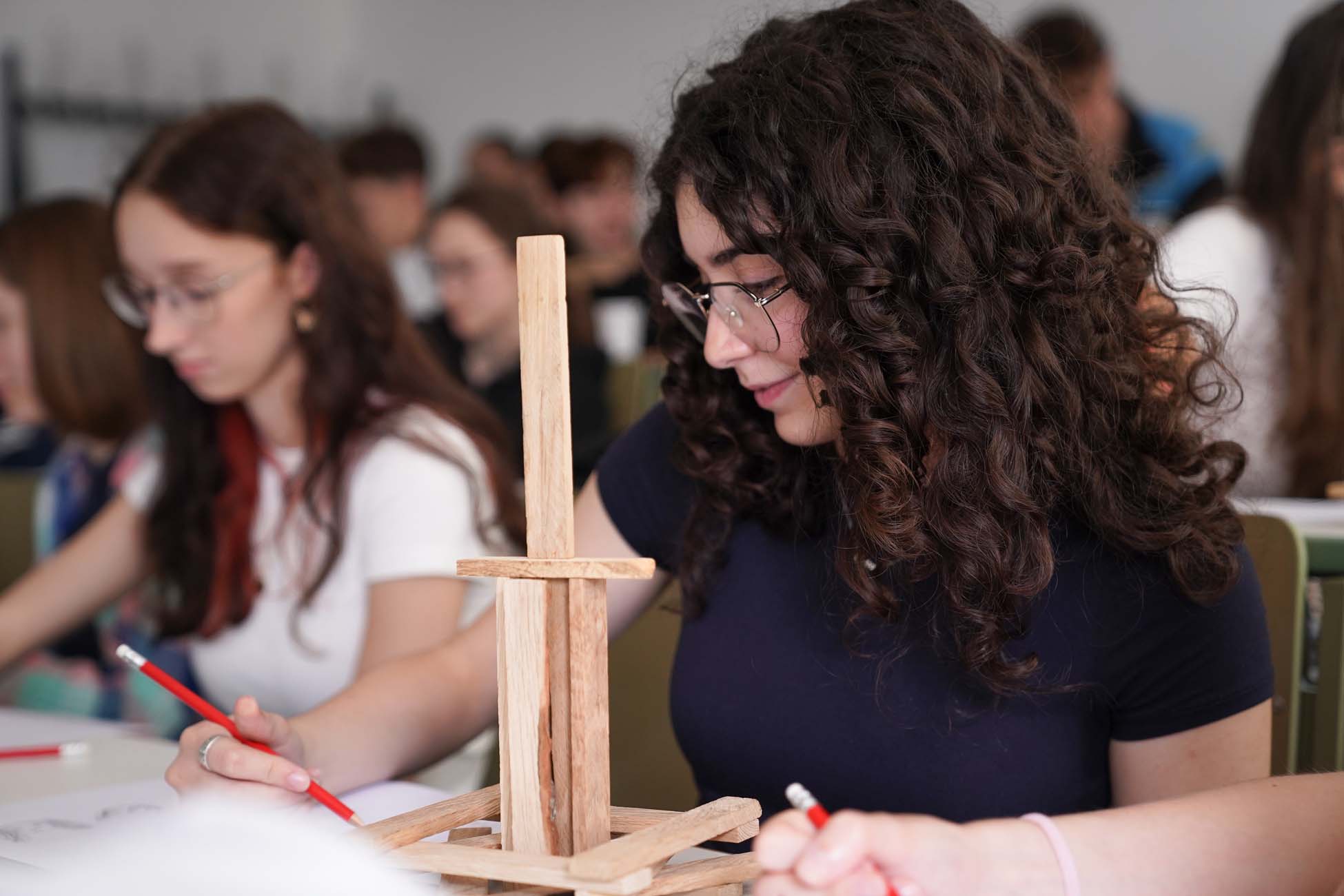 Eine Jugendliche mit lockigem Haar und Brille studiert an ihrem Schreibtisch eine Holzstruktur mit einem roten Bleistift, während sie von anderen Studierenden der Hochschule Coburg im Klassenraum umgeben ist und gemeinsam erkunden lernt. at Hochschule Coburg