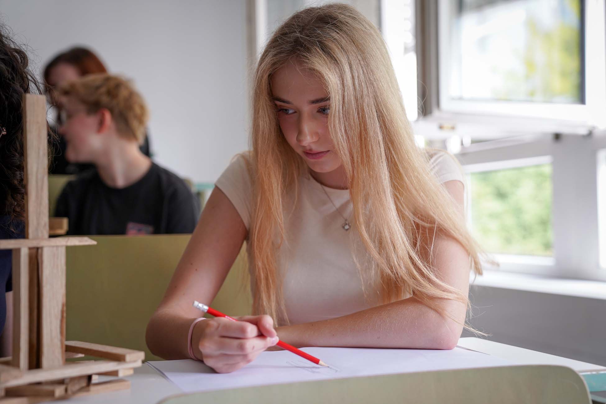 Eine junge Frau mit langen blonden Haaren sitzt an einem Schreibtisch am Fenster und zeichnet konzentriert mit einem roten Bleistift auf weißem Papier - eine typische Szene unter Jugendlichen an der Hochschule Coburg. at Hochschule Coburg