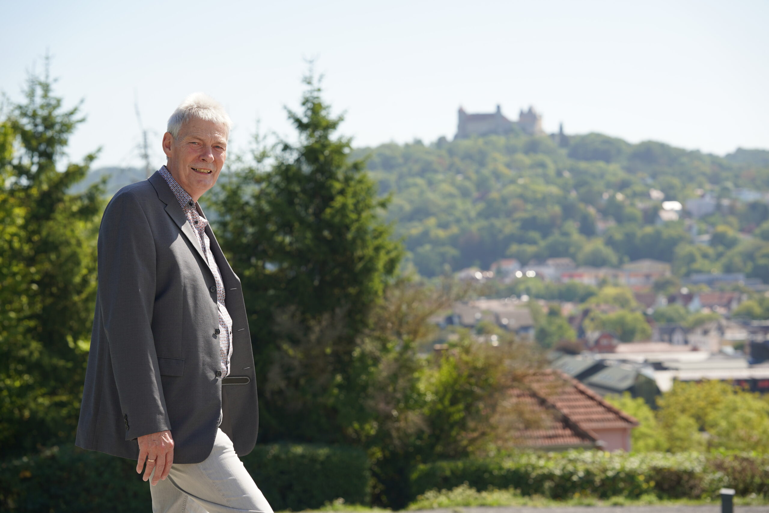 Prof. Dr. Vogt, im Anzug, lächelt bei seiner Verabschiedung im Freien. Unter dem klaren Himmel sind Bäume, Dächer und in der Ferne eine Burg auf einem Hügel zu sehen - eine passende Szene zum Abschluss seiner Professur. at Hochschule Coburg