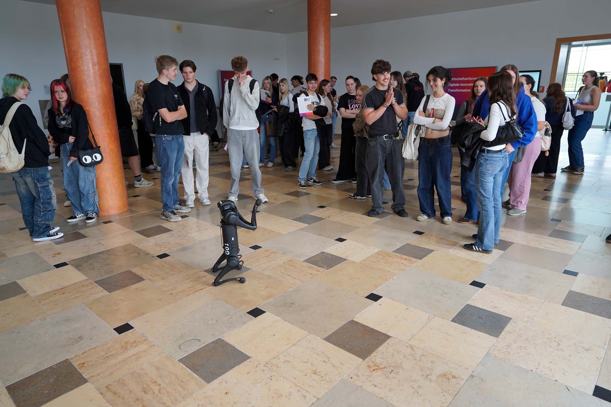 Eine Gruppe Jugendlicher steht in einem Foyer im Halbkreis versammelt und beobachtet, wie ein kleiner, schwarzer Roboterhund den Fliesenboden vor ihnen erkundet. at Hochschule Coburg