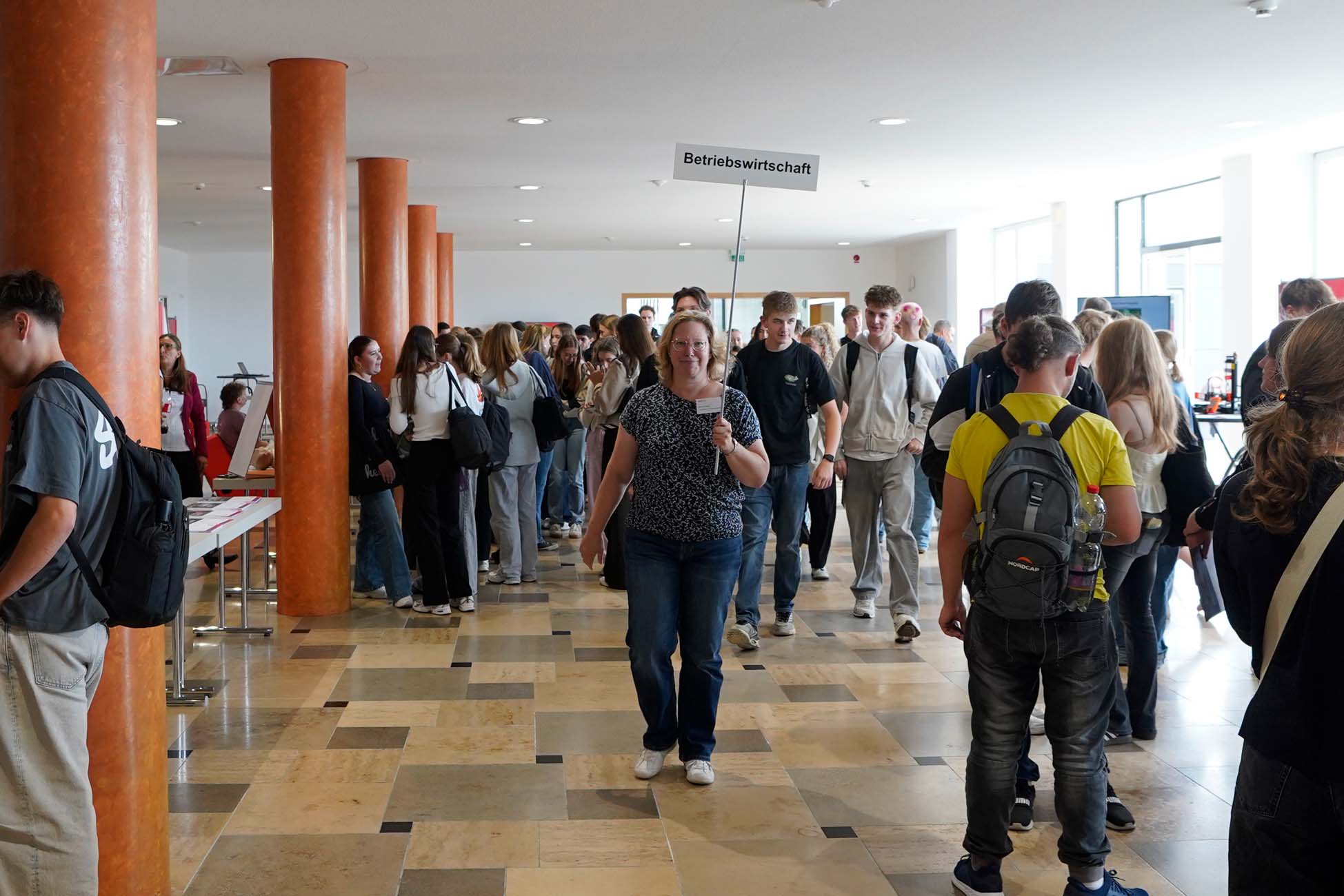 Eine Gruppe von Jugendlichen steht und geht in einem hellen Flur der Hochschule Coburg. Eine Frau in der Mitte hält ein Schild mit der Aufschrift "Betriebswirtschaft". Einige tragen Rucksäcke und unterhalten sich, andere erkunden die Umgebung. at Hochschule Coburg