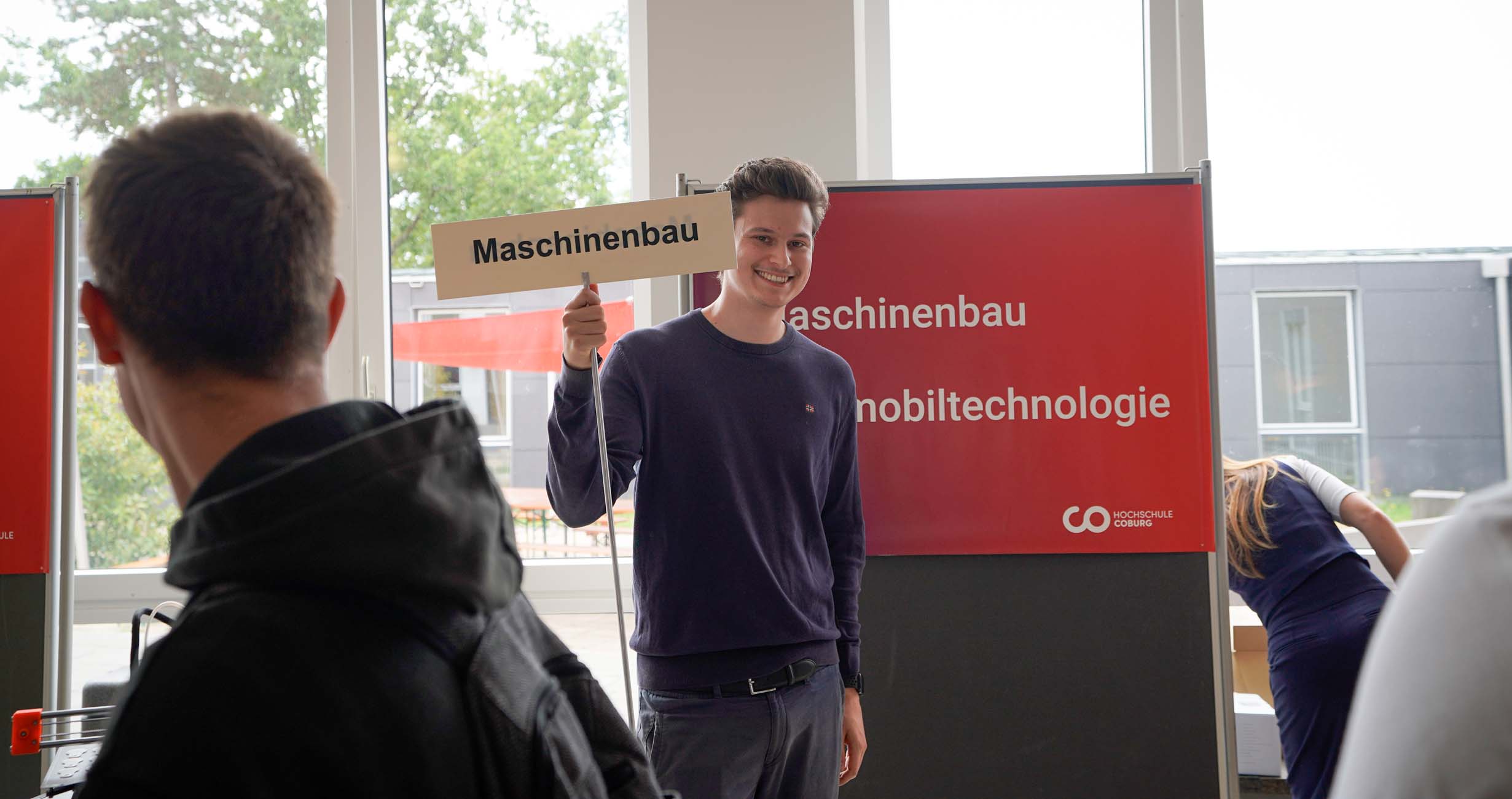 Ein junger Mann steht lächelnd drinnen und hält ein "Maschinenbau"-Schild. Hinter ihm erkunden Jugendliche und andere Besucher die roten Infotafeln bei einer Veranstaltung an der Hochschule Coburg. at Hochschule Coburg