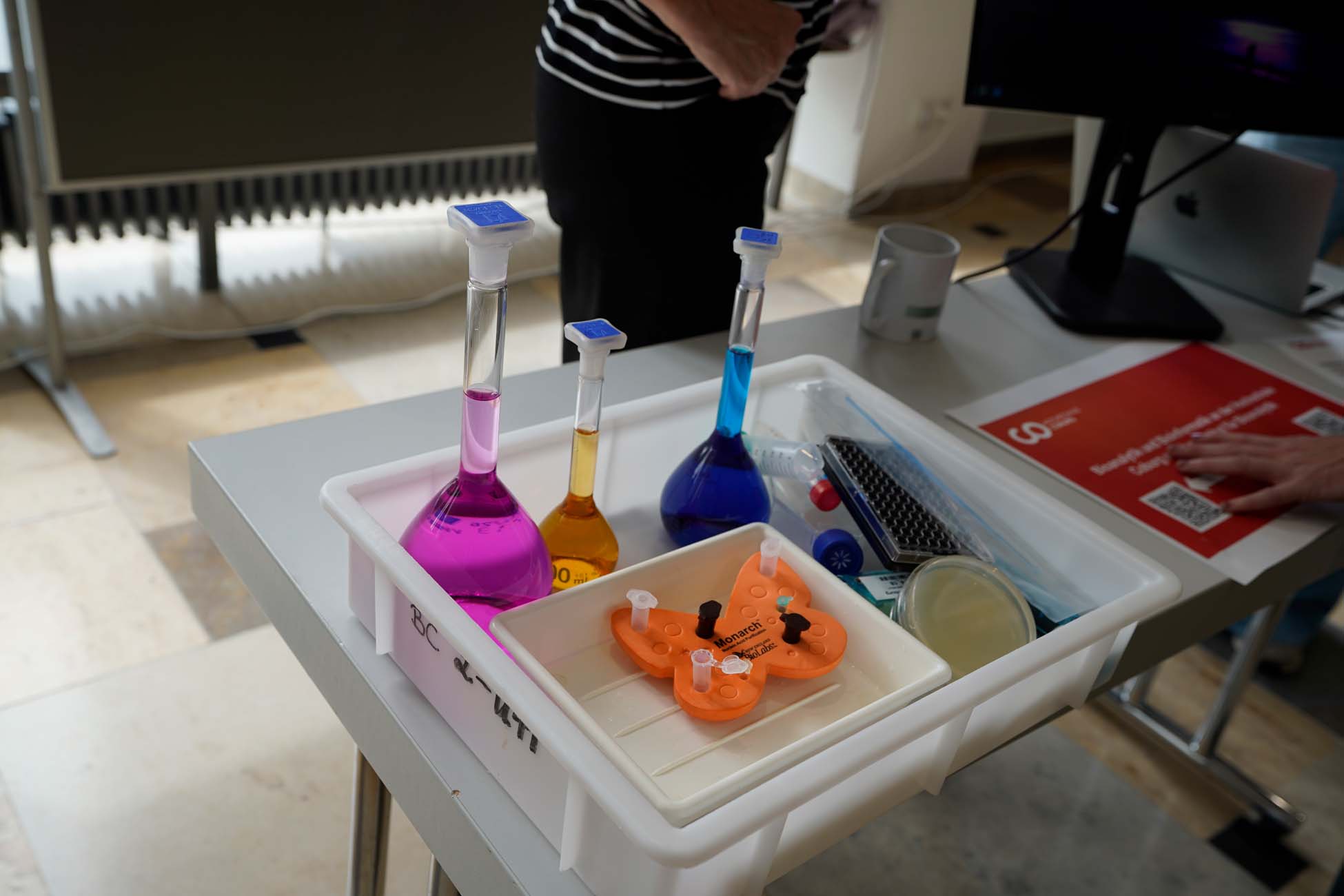 Ein Plastiktablett auf einem Tisch enthält drei Fläschchen mit lila, gelben und blauen Flüssigkeiten, Petrischalen und ein orangefarbenes schmetterlingsförmiges Objekt, das Jugendliche der Hochschule Coburg erkunden; im Hintergrund sind eine Person und ein Computer zu sehen. at Hochschule Coburg