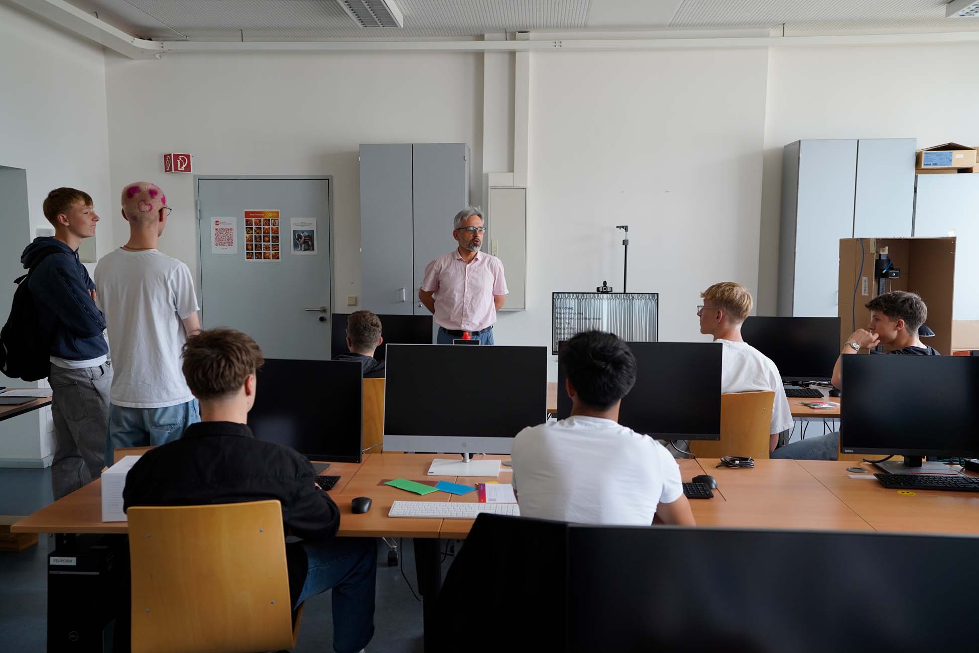 Ein Lehrer steht vorne in einem Klassenzimmer der Hochschule Coburg und spricht zu Jugendlichen, die an Tischen mit Computermonitoren sitzen; drei Schüler stehen in der Nähe der Tür, während vier sitzen und den Unterricht aufmerksam erkunden. at Hochschule Coburg