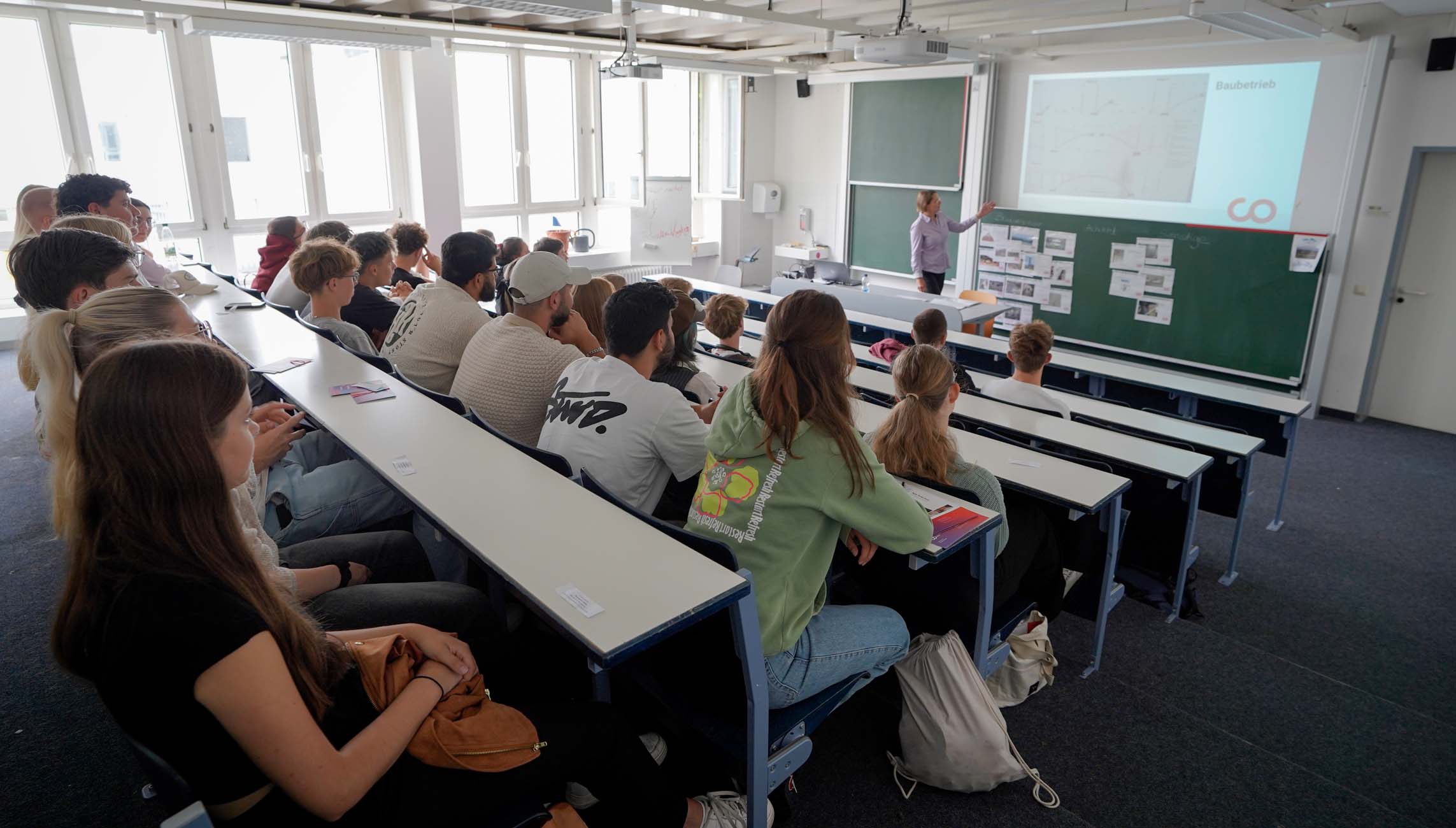 Ein Klassenzimmer voller Jugendlicher sitzt in gestaffelten Reihen vor einem Dozenten der Hochschule Coburg, der einen Vortrag hält. Der Dozent steht vor einem Beamer und einer Kreidetafel, während die Studenten unter hellen Fenstern mit natürlichem Licht neue Ideen erkunden. at Hochschule Coburg