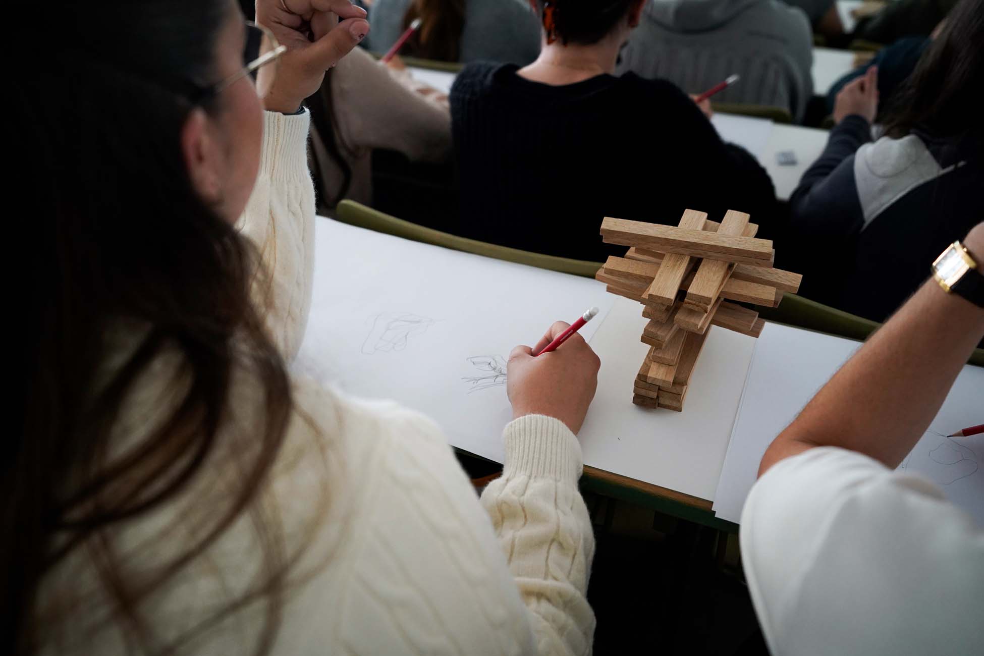 Ein Jugendlicher hält einen Rotstift in der Hand und skizziert auf Papier neben einem geometrischen Holzmodell in einem Klassenraum der Hochschule Coburg, umgeben von anderen, die ebenfalls ihre Kreativität zeichnen und erkunden. at Hochschule Coburg
