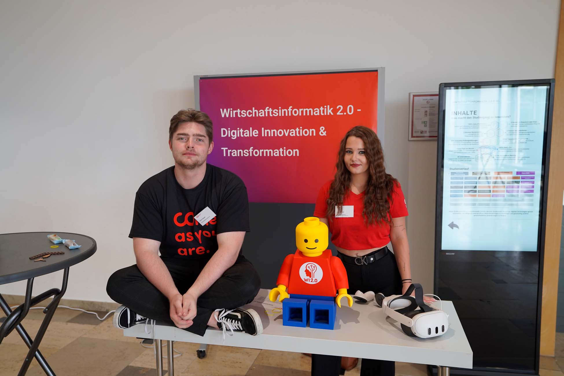 Zwei Jugendliche erkunden einen Stand mit dem Schild "Wirtschaftsinformatik 2.0 - Digitale Innovation & Transformation" an der Hochschule Coburg. Auf dem Tisch liegen eine LEGO-Figur und ein VR-Headset; einer sitzt im Schneidersitz, der andere steht. at Hochschule Coburg