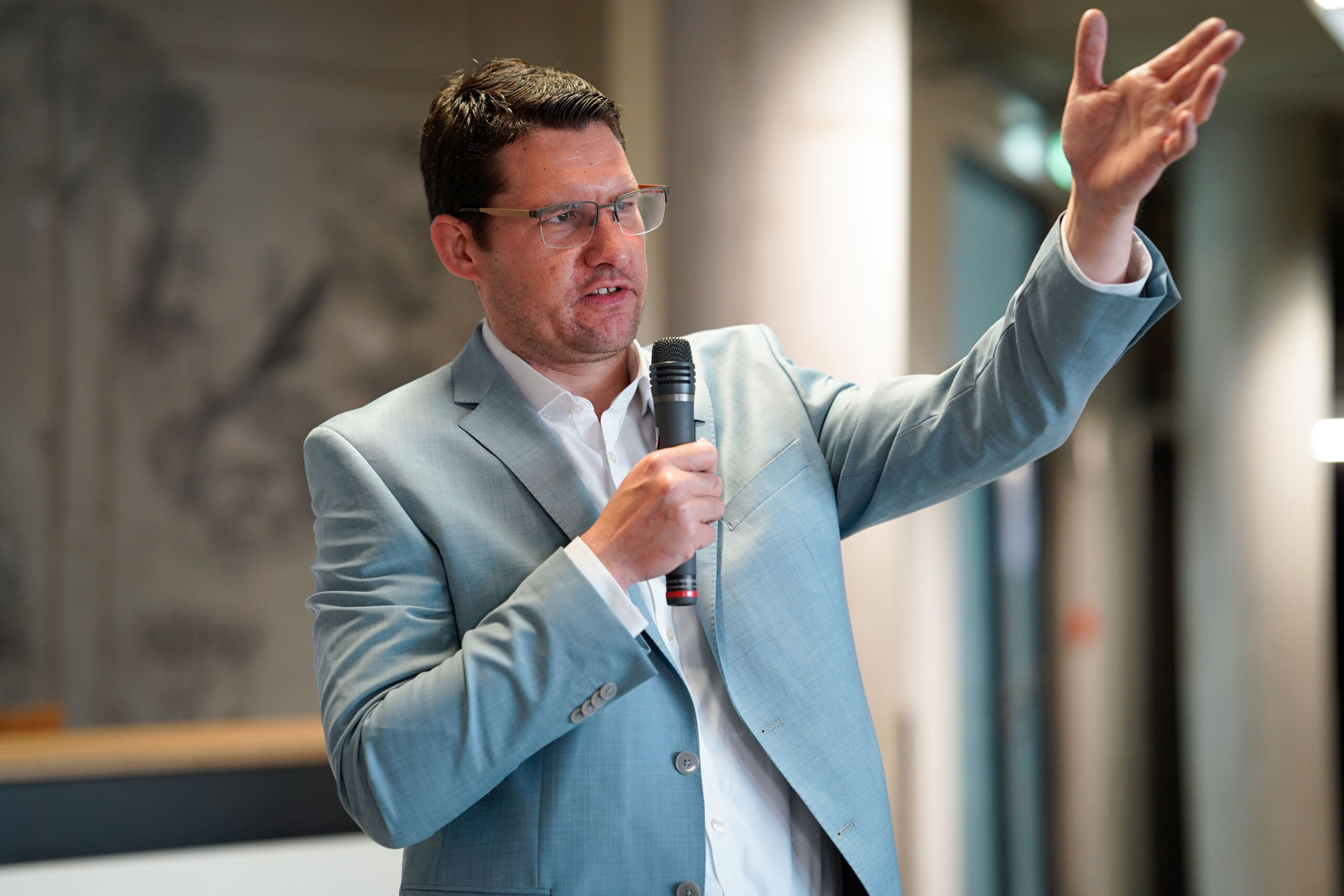 Ein Mann im hellblauen Anzug und mit Brille spricht in ein Mikrofon und gestikuliert mit der erhobenen rechten Hand, während er in einem Innenraum einer modernen Werkschau mit Schwerpunkt auf 3D-Druck-Innovationen präsentiert. at Hochschule Coburg