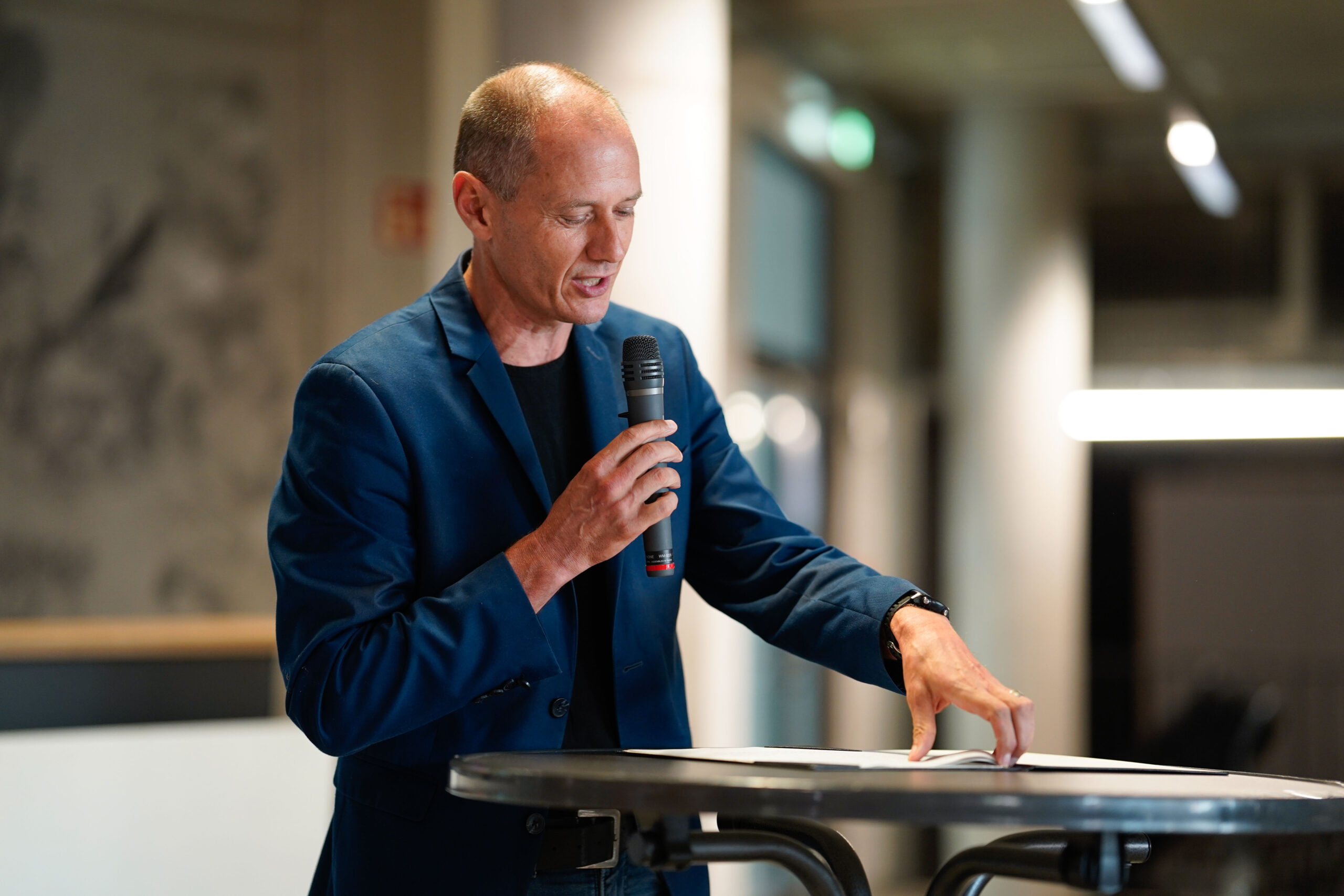 Ein Mann in einem blauen Blazer spricht in ein Mikrofon, während er auf ein Dokument auf einem runden Tisch zeigt. Er diskutiert über Quartiersumbau in einem modernen, gut beleuchteten Innenraum während der Werkschau. at Hochschule Coburg