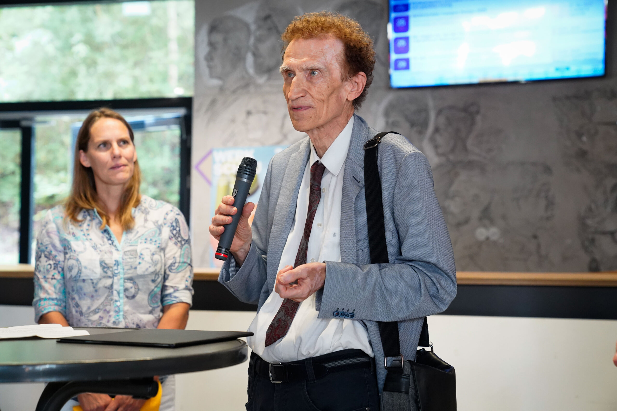 Ein Mann im Anzug hält ein Mikrofon und spricht neben einer Frau in einem gemusterten Hemd während der Quartiersumbau Werkschau. Sie befinden sich in einem Innenraum in der Nähe eines Tisches, mit einer Leinwand und einem Fenster im Hintergrund. at Hochschule Coburg