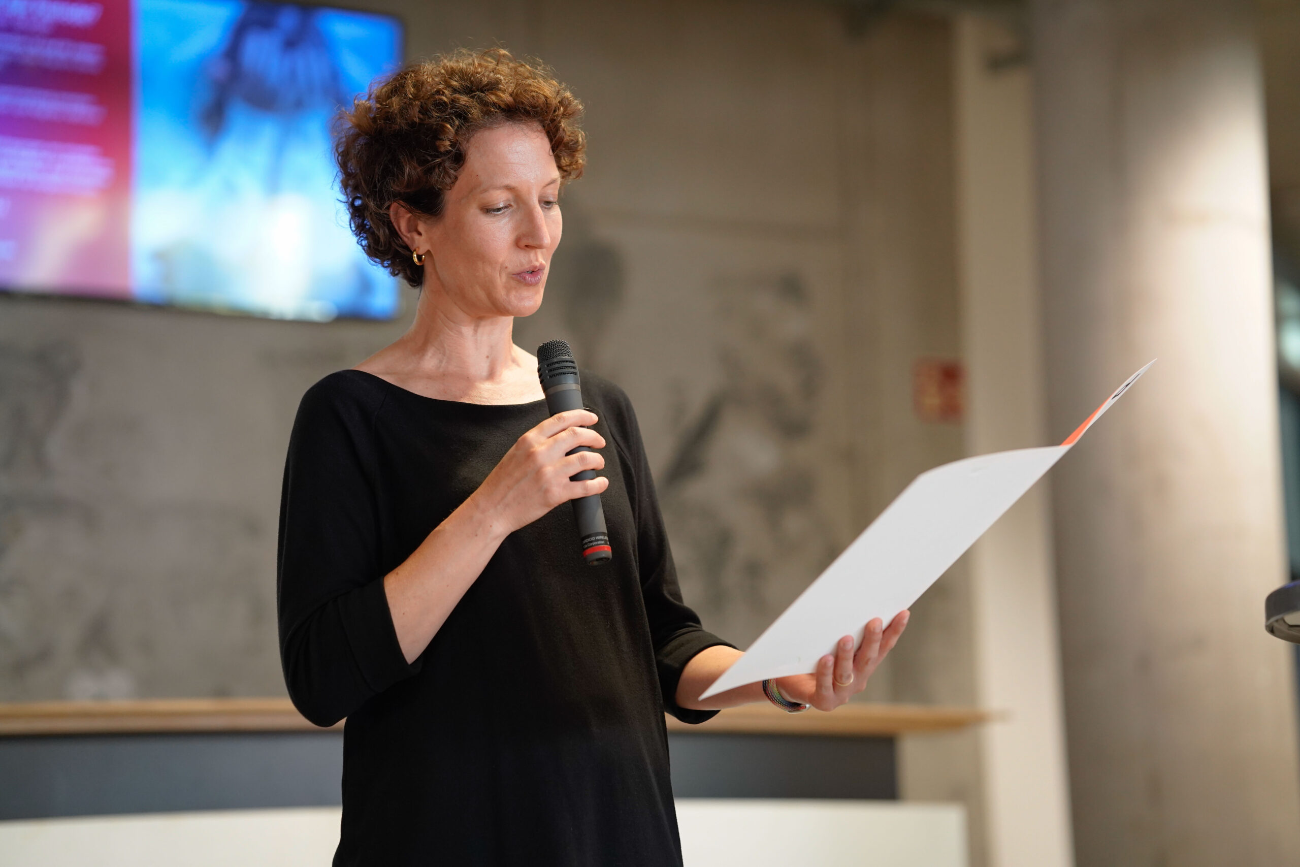 Eine Frau mit kurzen lockigen Haaren, die ein schwarzes Oberteil trägt, hält ein Mikrofon in der Hand und liest von einem Blatt Papier ab. Sie spricht in einem Innenraum vor einem unscharfen Hintergrund - vielleicht ein Tabubruch oder eine Diskussion über Abschlussarbeiten bei einer bdia-Veranstaltung. at Hochschule Coburg