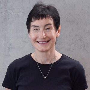 HüttlAnnerose Annerose Hüttl, eine lächelnde Frau mit kurzen dunklen Haaren, die ein schwarzes T-Shirt und eine silberne Halskette trägt, steht vor einer hellgrauen, strukturierten Wand. at Hochschule Coburg