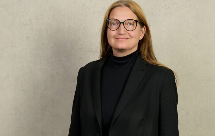 Eine Person mit langen hellbraunen Haaren und Brille trägt einen schwarzen Blazer und einen schwarzen Rollkragenpullover und steht vor einer schlichten hellen Wand für ein BR-Interview mit Prof. Meißner über offene Placebos. at Hochschule Coburg